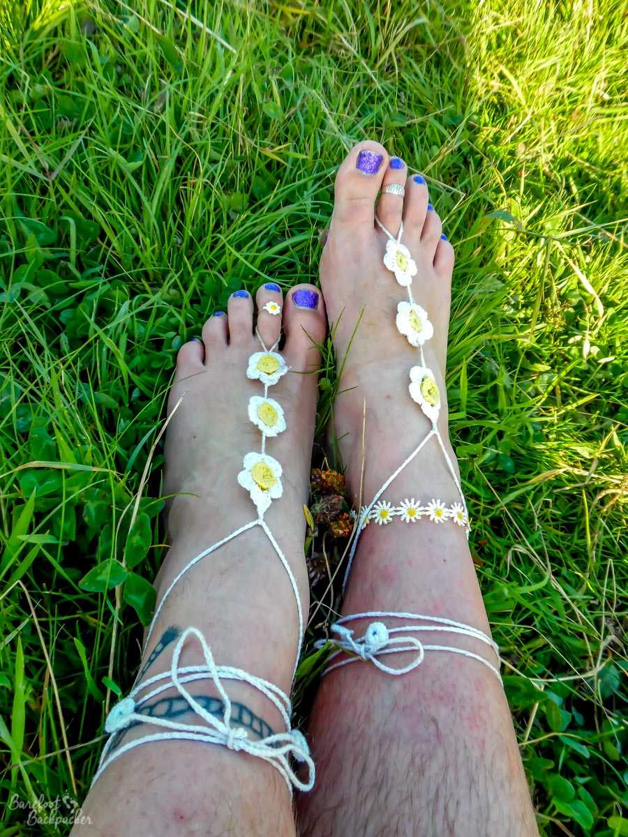 Traditional barefoot sandals being worn. Two feet, otherwise bare, lie in the grass. The sandals are made up of a length of white string tied round the ankle and looped around the toe next to the big toe. As they trail along the the top of the foot, there are three 'daisies' woven into them.
