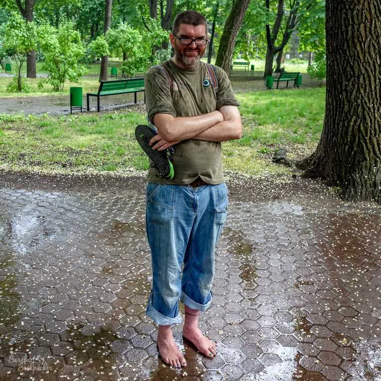 I'm standing in a city park, on some hexagonal paving, in front of trees and grass. It's raining. I'm looking grumpy and damp. I'm wearing a sodden long-sleeved t-shirt, cuffed jeans, and am barefoot - I'm holding my sandals in my hand.