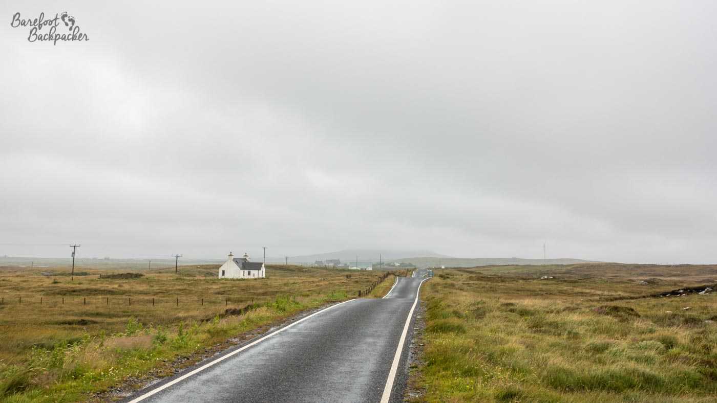 A remote, empty, road disappears straight into the distance, below a grey, moody, sky. One building lies on the left side; the world otherwise is a bleak, remote, coastal moorland.  This is North Uist, in the Outer Hebrides of Scotland.