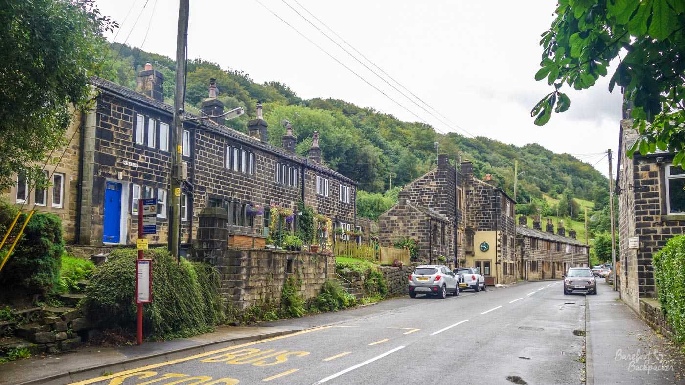 A street in the Cragg Vale, Calderdale Borough, West Yorkshire. It's a fairly grey day, just after a rain shower. The view is of a road lined with old fashioned stone cottages, in front of some fairly small but steep hills in the background.