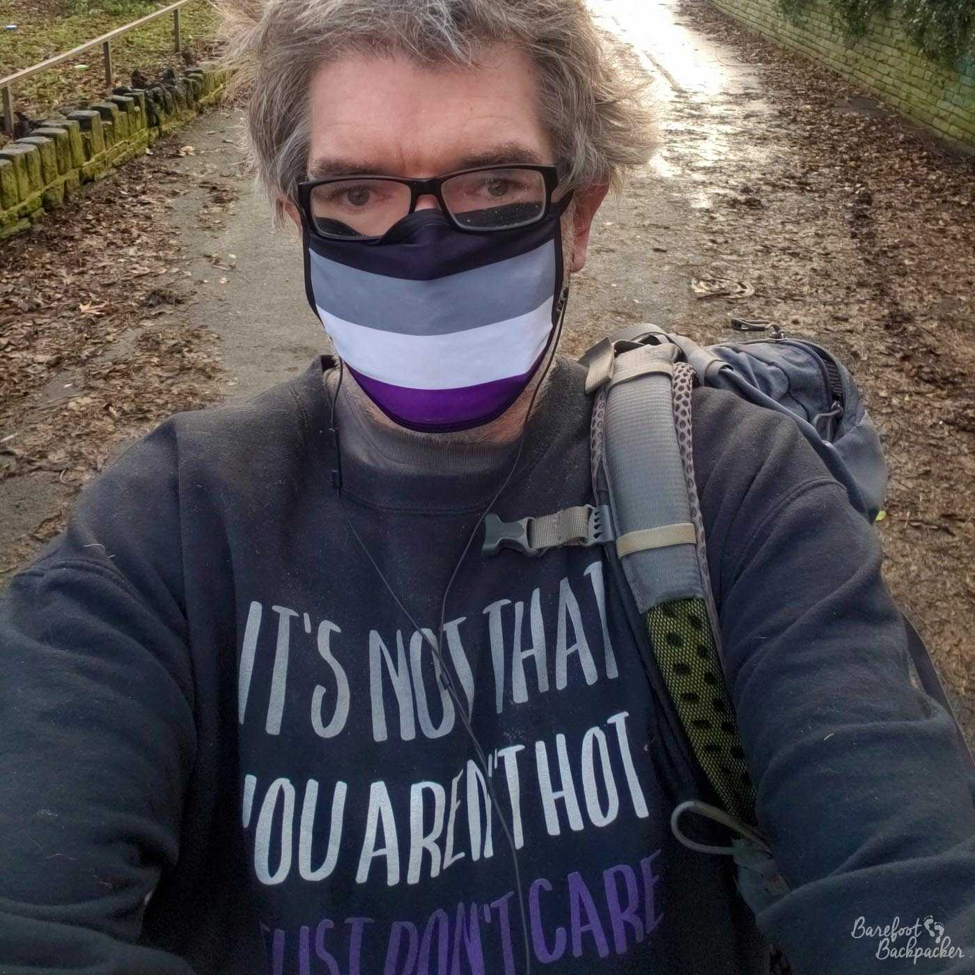 Man (again, that's tbd) standing on a pedestrianised street on a hill. The backpack is still there, over one shoulder and empty, but this time they're wearing a black sweatshirt with the caption 'it's not that you're not hot, I just don't care' in white, grey, and purple. They're also wearing a face-mask with black, grey, white, and purple stripes.