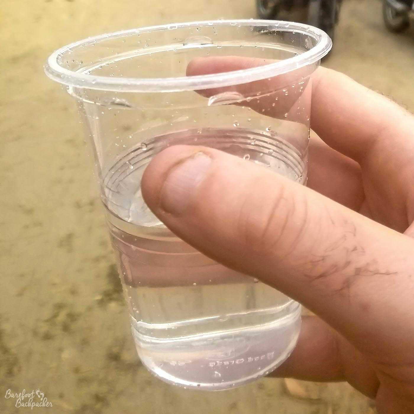 A hand holding a plastic cup of clear, transparent, water-like liquid. It is not water. It is very far from being water.