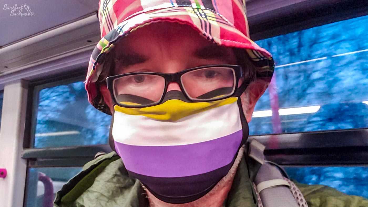 A face on a tram, mostly hidden by them wearing a face-mask in the colours of the non-binary pride flag - yellow, white, purple, black. They're also wearing a plaid, mainly red/white, hat