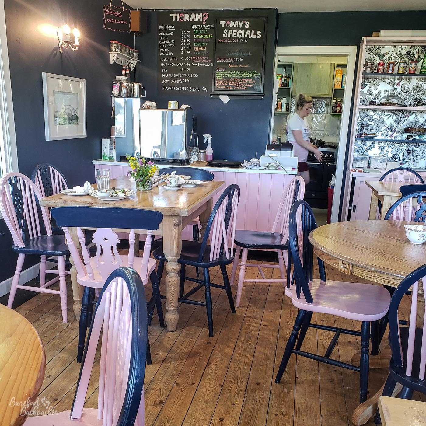 Inside the Tioram Cafe in Acharacle. Black chairs with pink seats, a wooden floors, pink wooden serving counter, wooden tables (light brown). A chalkboard behind the counter with the menu on it. Door to the kitchen open, a woman can be seen making stuff in it. To the right of the door is a cupboard with shelving, the back of the board is what looks like drawn flowers on wallpaper. The walls are plain darkish blue.