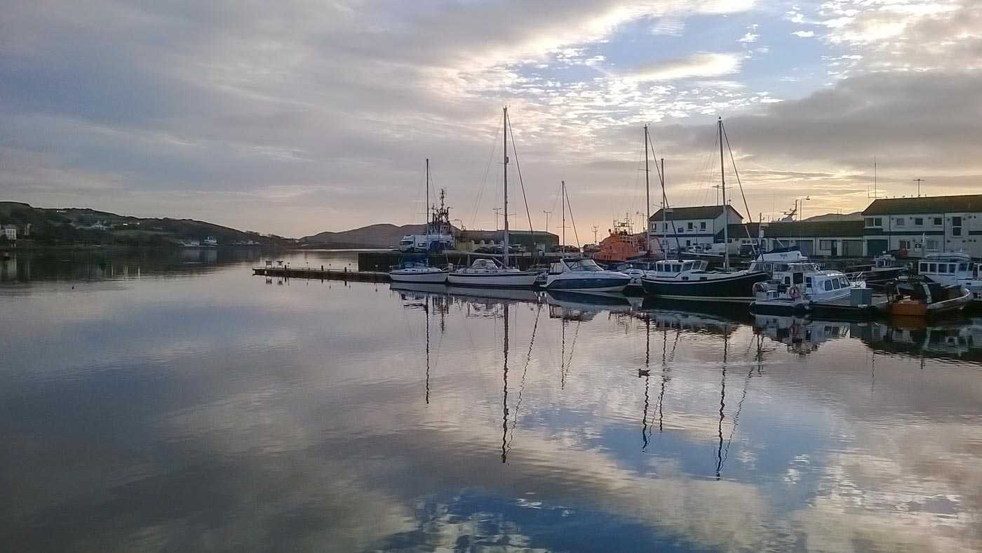 A series of yachts lie on the smooth surface of Campbeltown marina, their masts reflecting in the water. Some white buildings lie behind them, but otherwise the setting looks very serene.