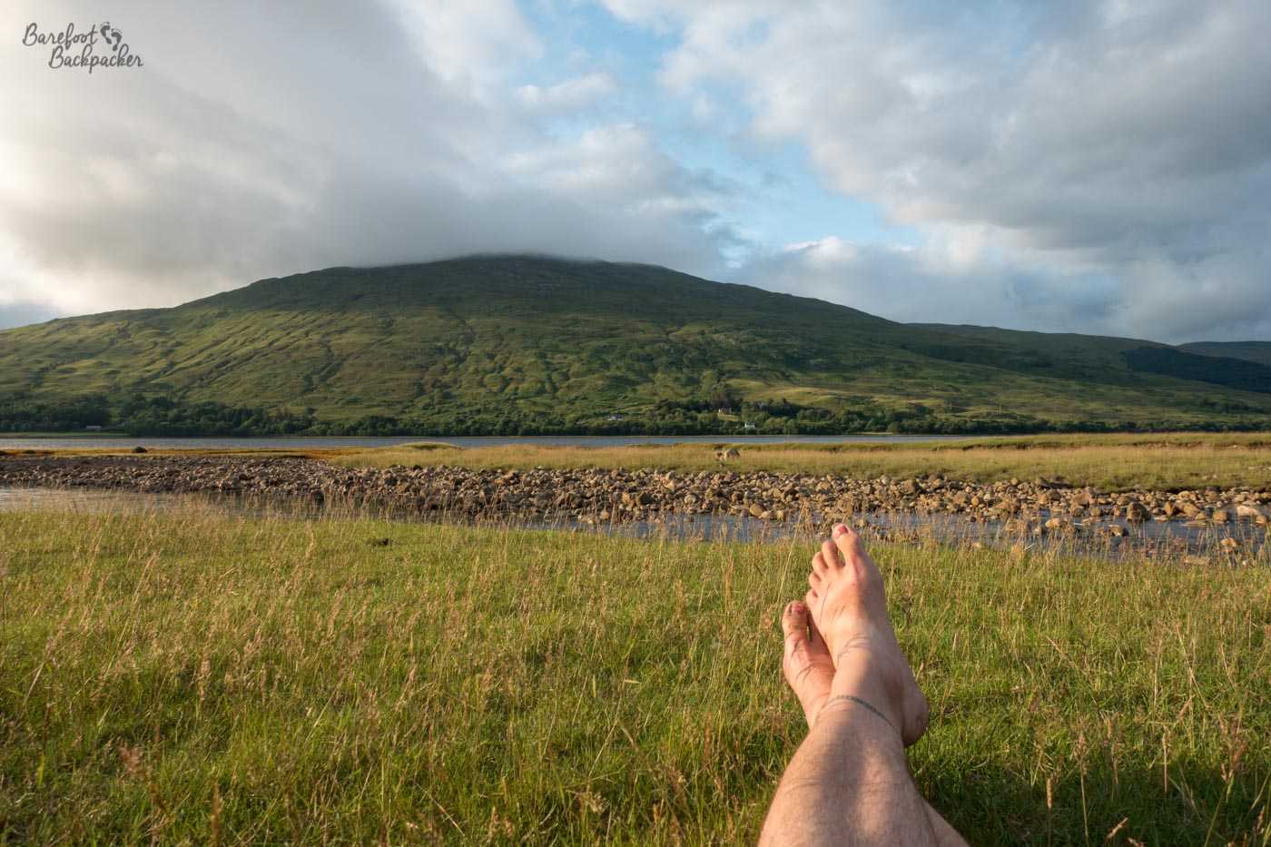 Low-lying grasses, a stream with a  rocky pebbly bank, then more grasses, then a flat loch. The upper half of the image is dominated by a green hill rising beyond the far side of the loch. On the lower right  of the shot are two legs resting flat on the grass, ending in a pair of bare feet, crossed at the ankles. The sun is shining, giving a late evening brightness to proceedings.