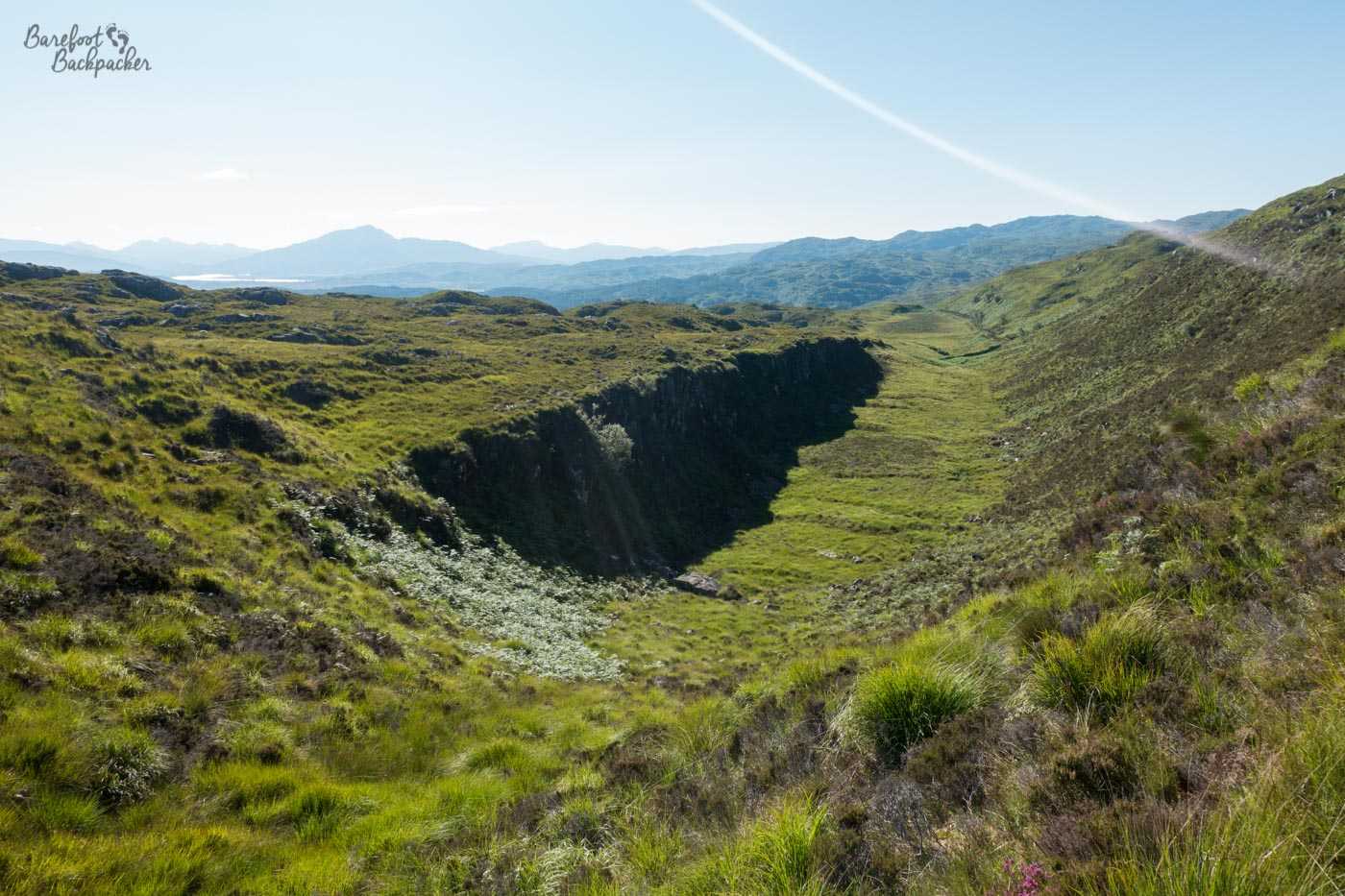 What appears to be a small dry chasm, green and thick with grasses. The right side is a steep but gradual slope up to a ridge where there is a path. The left side is a sheer cliff up to a ridge covered in shrubs and grasses. Misty blue hills in the background, bright sunshine otherwise.