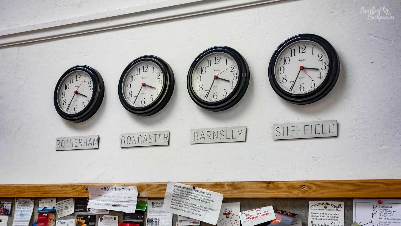 Four clocks on a wall in a cafe. They are located above a shelf of information leaflets and adverts. The clocks are similar to those you find in multinational office buildings, which show the times in different cities around the world. These four depict the time in the four main towns of South Yorkshire. Oddly, they're not quite aligned, a couple of places are implied to be in a time-zone of GMT+17 seconds.