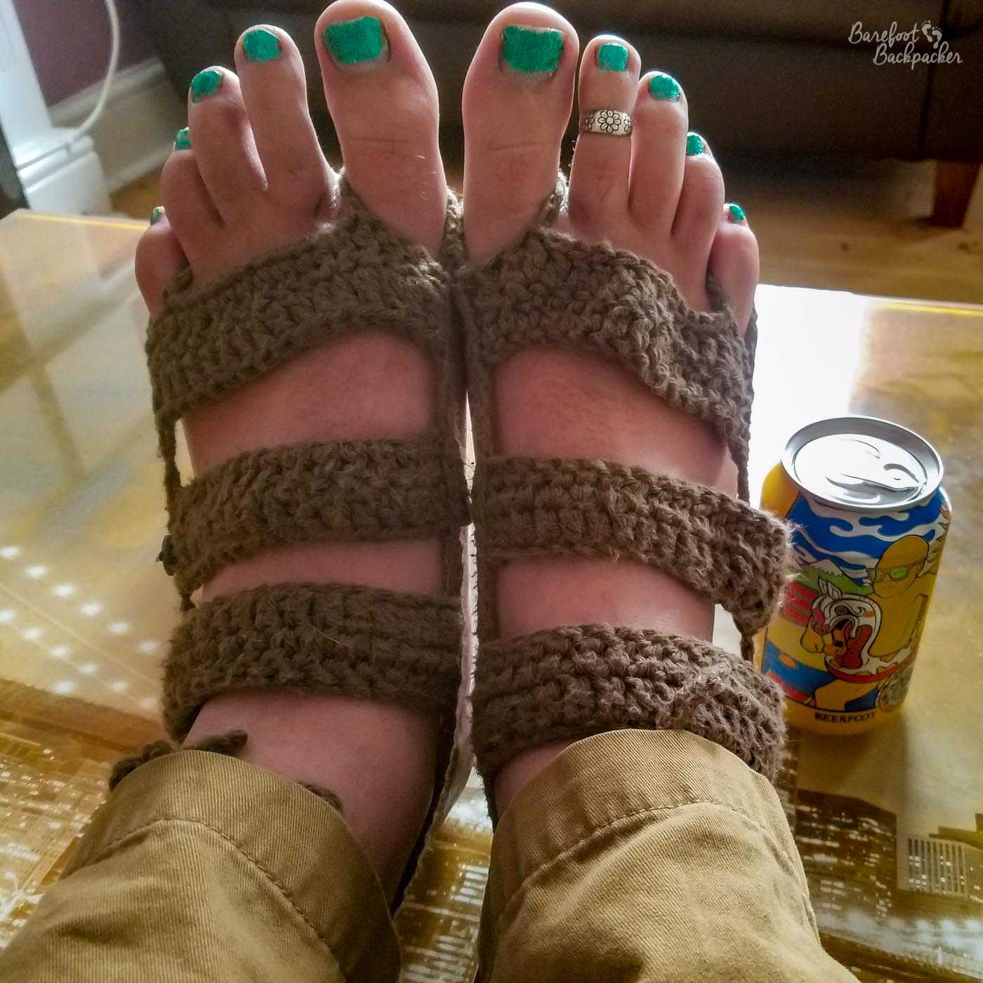 The new barefoot sandals. Three thick brown crocheted straps across the width of the top of each foot, from the toes to the ankle. Loops of thread go round both the big toes and the little toes. Toenails are green and sparkly, the feet are resting on a glass table, and to the right of the right foot is a can of beer called 'Beerfoot'.