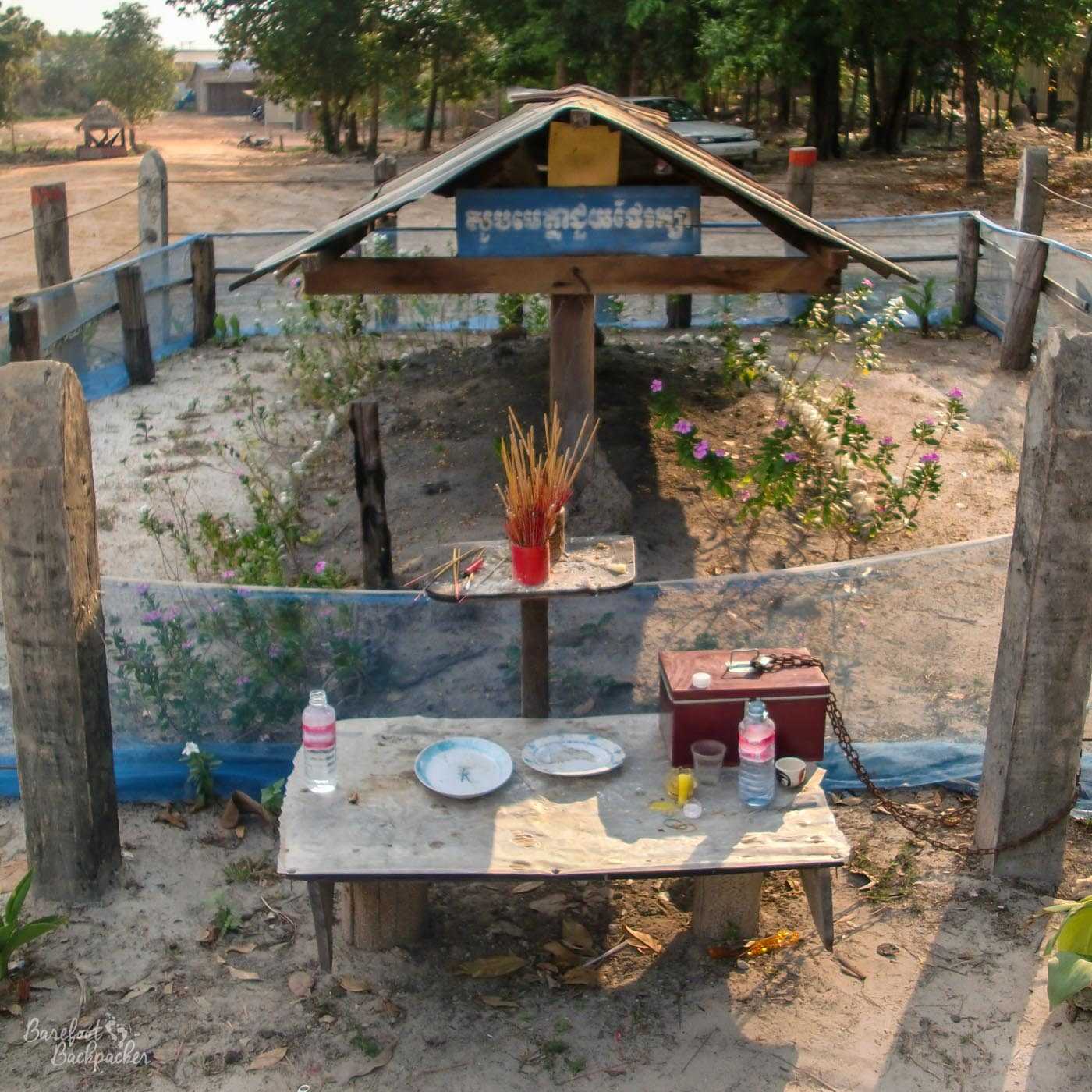 Pol Pot's cremation and burial site, in Anlong Veng, at the border with Thailand. It's a mound of soil under a wooden construction that has the feel of a large bird table. There are purple flowers with long stems growing on it. It is surrounded by a thin chicken wire fence. In front of the fence, at one end of the gravesite, is a makeshift table with plates, plastic water bottles, a metal box chained to a concrete fencepost, and a couple of jars of incense sticks.
