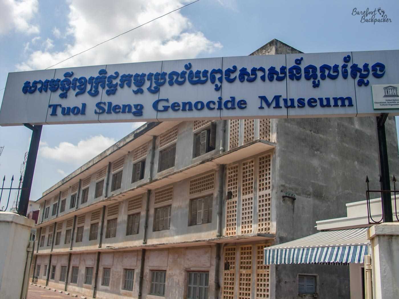 Signpost for the Tuol Sleng Genocide Museum. In the background is a concrete block of a three-storey building, with shuttered windows and ventilation bricks.