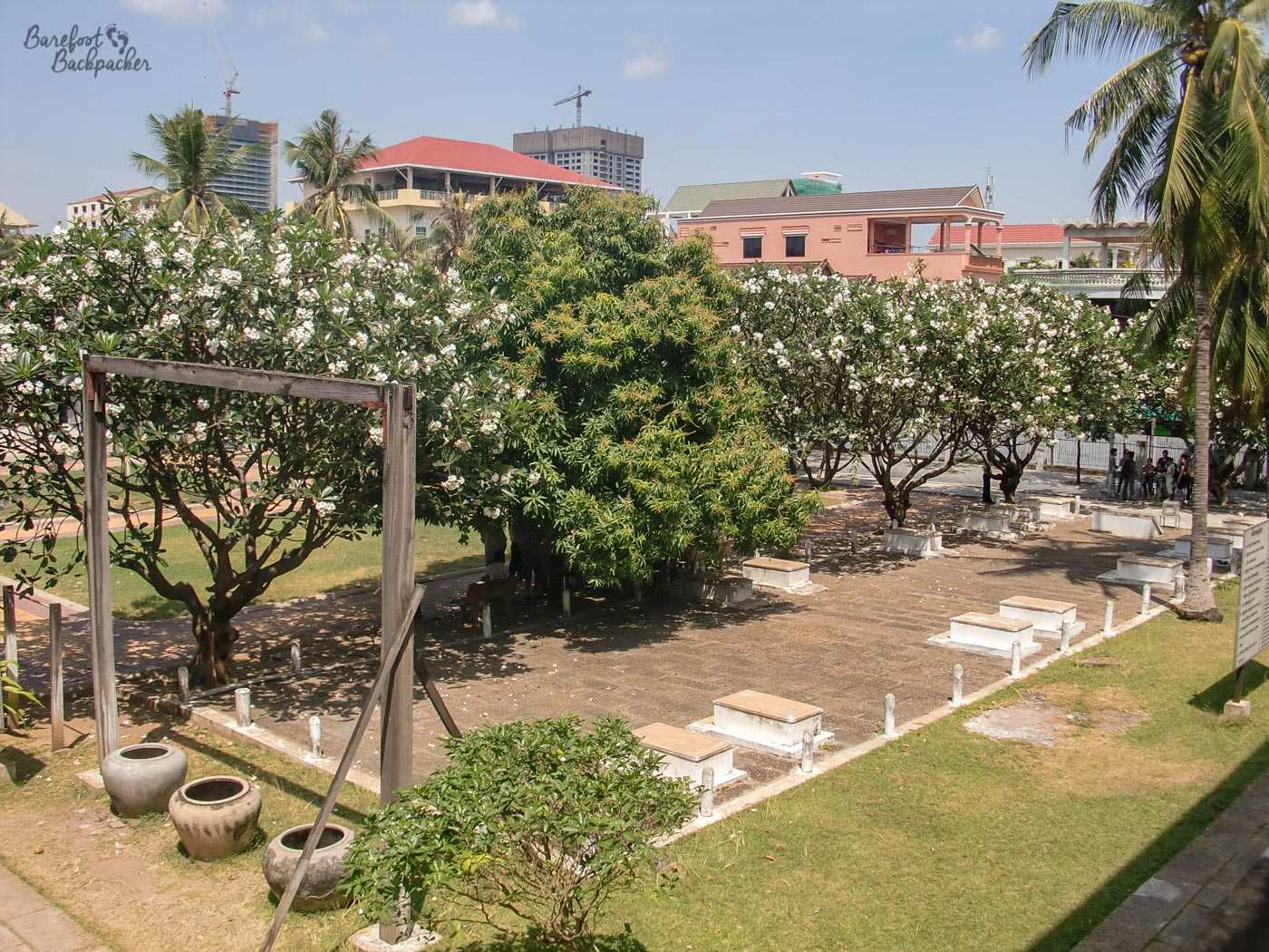 View out of one of the upper floor classroom windows onto what was the playground. There is a yard lined with coffins, symbolic representations of those who died. On the left, at the near end of the yard, is a wooden frame, with ceramic pots on the floor under the cross-beam. In Khmer Rouge days people would be hanged upside down by the ankles with their heads in the buckets. The buckets would generally have been filled with excrement.