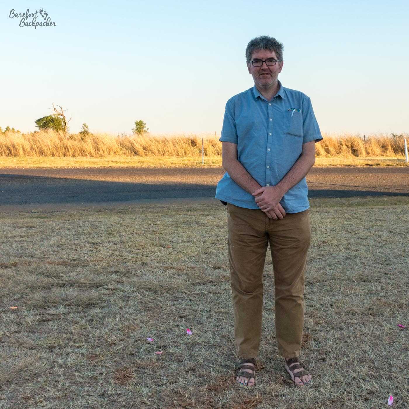 Standing in a field in Queensland, Australia (Henley Brook). The man wears a blue short-sleeved buttoned shirt and light brown trousers, He is wearing the crochet barefoot sandals. The field is scrubland. Behind him is a road bathed in sunshine, and behind that a bright yellow 'hedge' of corn, set against a bright pale blue sky.