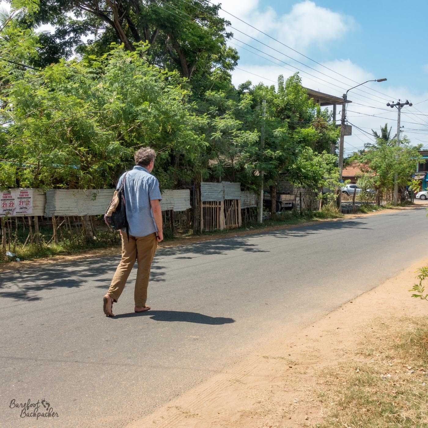 Walking down a street in Jaffna, Sri Lanka. Wearing a blue shirt, brown trousers, and the brown crochet barefoot sandals. However from this angle, it's clear, with the left foot up, on the toes, that the sole of the foot is bare and the sandals only cover the top. The road is dry tarmac, slightly dusty at the right edge, while the left side, behind the man, is lined with trees and wooden fences.