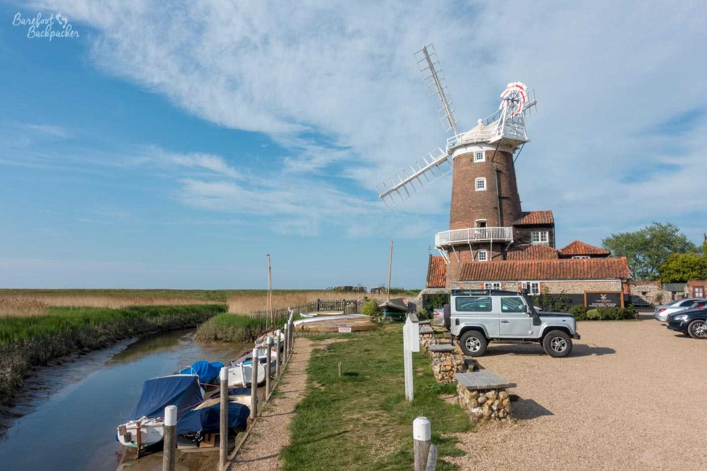 A small light brown brick windmill is on the right of the image, with white sails. On the left is a small muddy river. Behind are fields of long grasses. In front is a small car park with a 4x4 and a couple of cars in it. Between the car park and the river are a couple of boats. It is a blue sky with wispy light cloud.