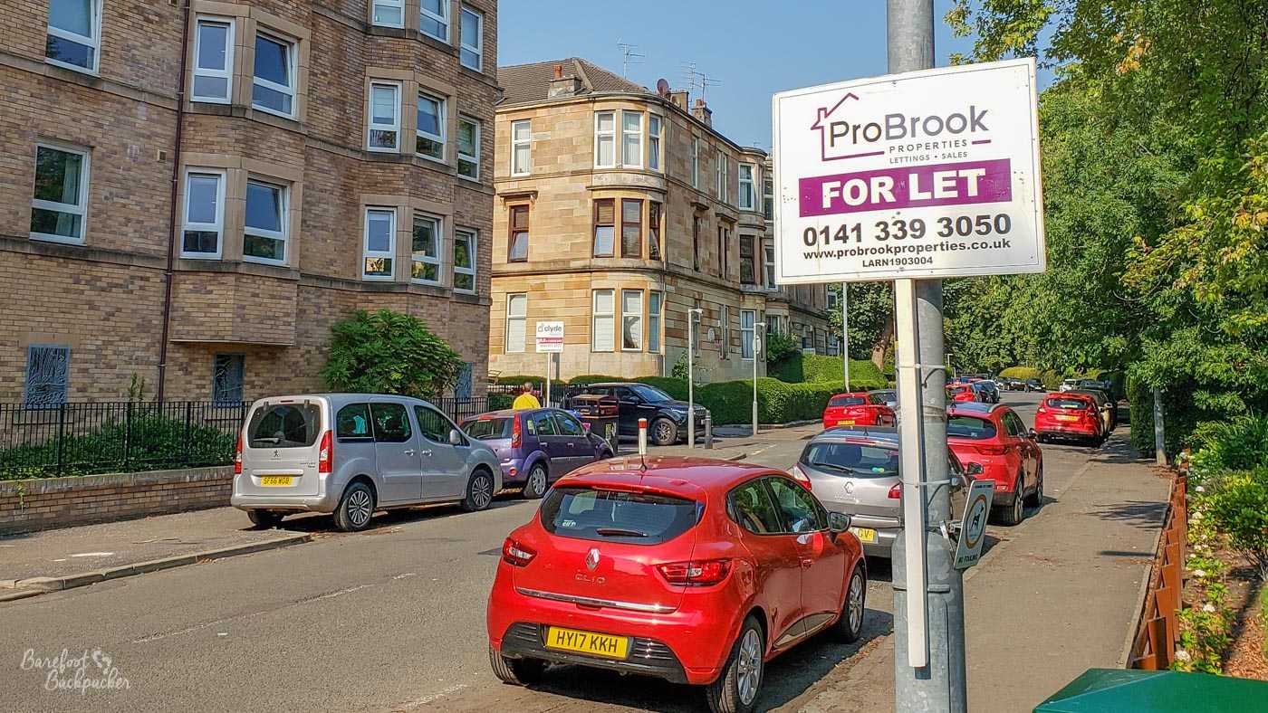 A city street, tenement blocks in the background, cars parked on the road in the middle. A couple of signposts ae visible, advertising properties available 'to let'