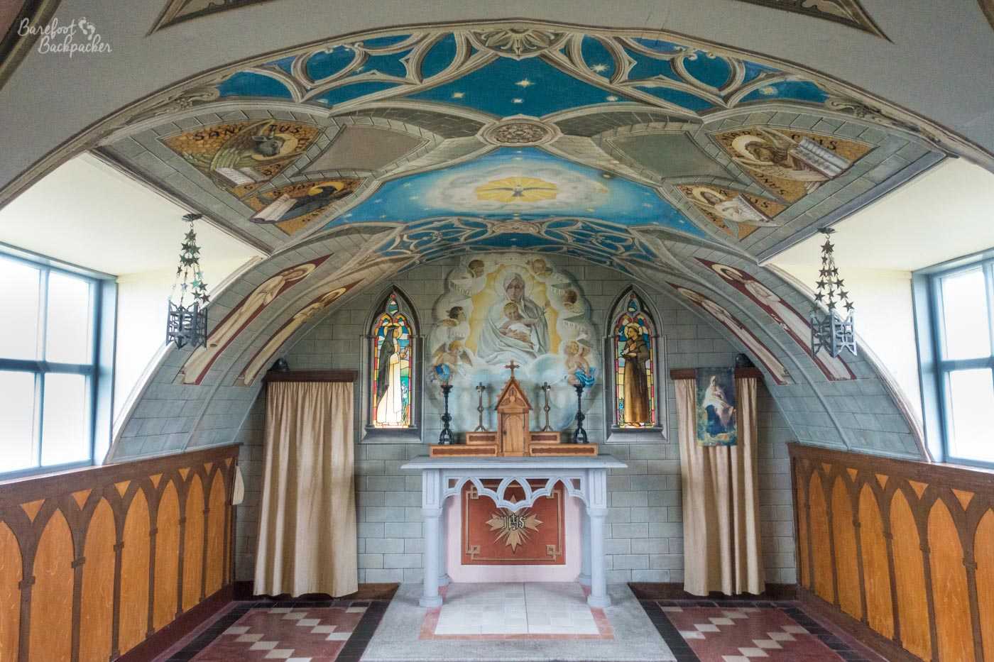 The inside of the Italian Chapel. The décor is mainly sky-blue and orange on white walls, with biblical frescoes on the ceiling and images of Jesus on the walls. In the centre of frame is a white altar with red decorations, also crucifixes and candlesticks, in front of a large Jesus/Mary fresco either side of two small stained glass windows.