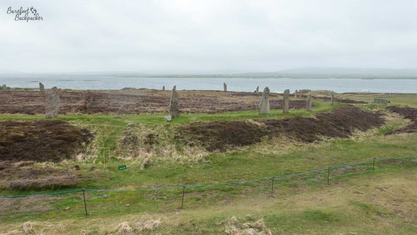 An overview shot of approximately 13 stones scattered around half a ring. The stones are clearly located on a plateau of grass, with an embankment behind them. Inside the stone ring it#s flat, but the land is covered with gorse or heather rather than grass. The shot is taken from atop a nearby mound. The loch is in the background.