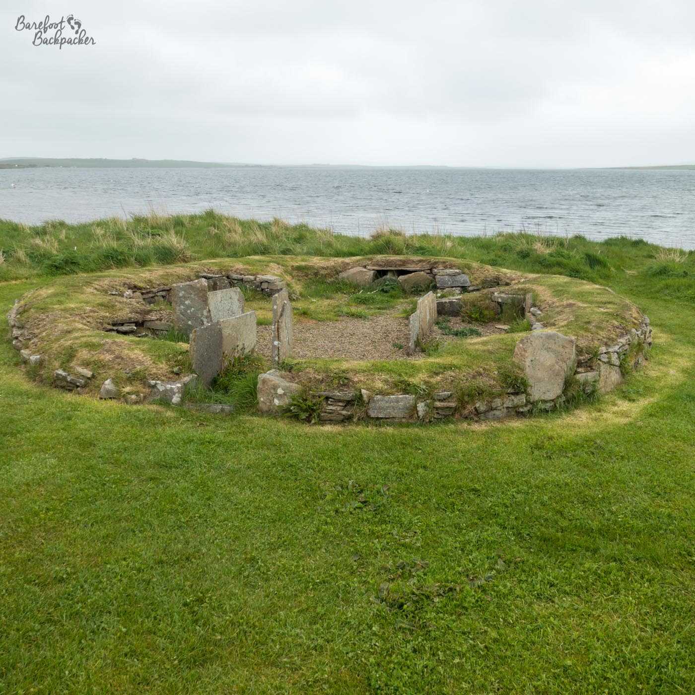 A slightly sunken stone ruin in a field – it's very much a ring shape but it's clearly a building – the entrance and the vague layout can be determined by the remains of the walls. The loch is close by in the background.