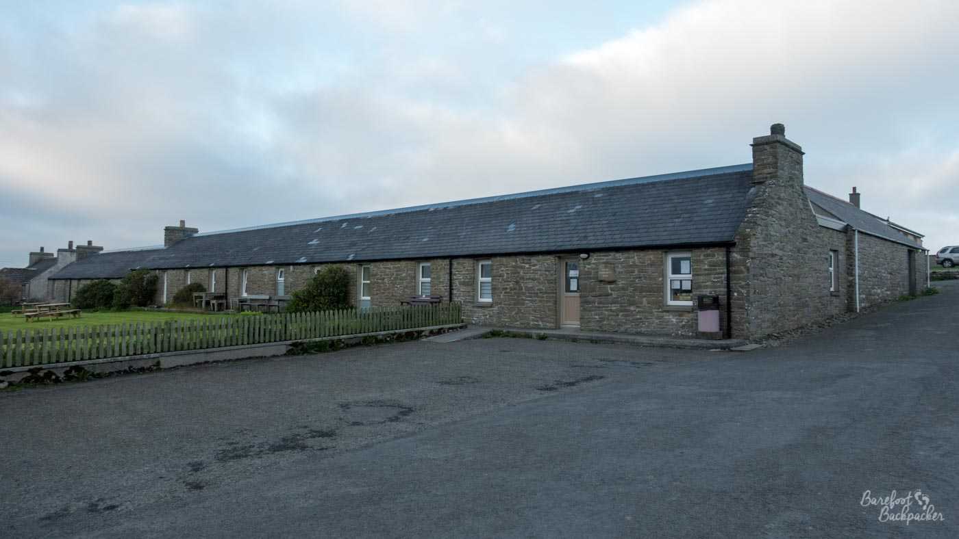 A long stone building stands half in a tarmac roadway and half next to a back garden. It's a one storey building. There's a bin on the wall at the right hand end, near a door with a couple of notices on it.