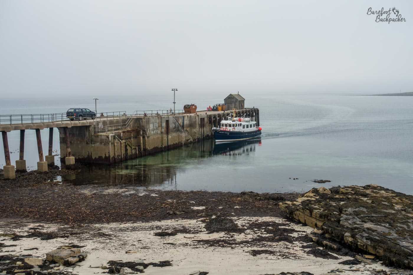 A small ferry slides up to a high stone quayside. There's a car on the quayside waiting to pick up or drop off passengers. In the foreground a beach slopes down to the water.