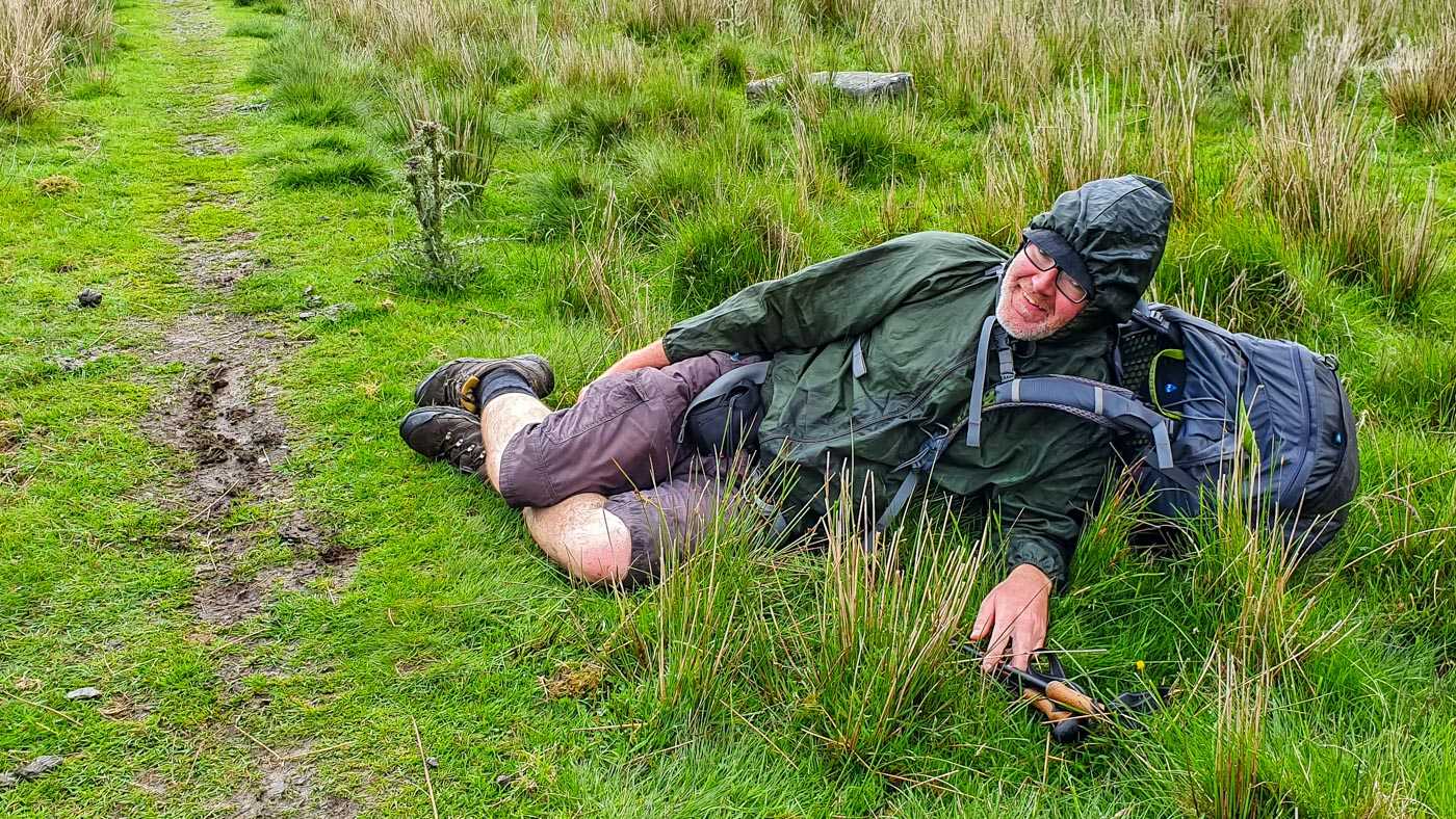 Muddy footpath through a field of grass. A man is lying on the grass just off the footpath. He's wearing a kagoul (hood up, covering his head), hiking cargo shorts, and walking boots, with a backpack on his back. He is laughing at the camera.