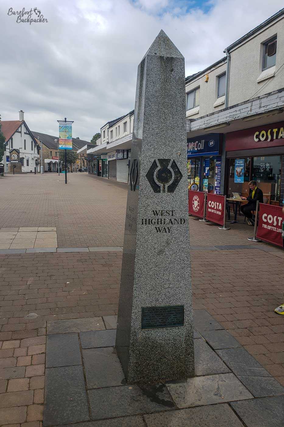 A stone obelisk, saying West Highland Way, stands on the pedestrianised high street, with shops to the right.