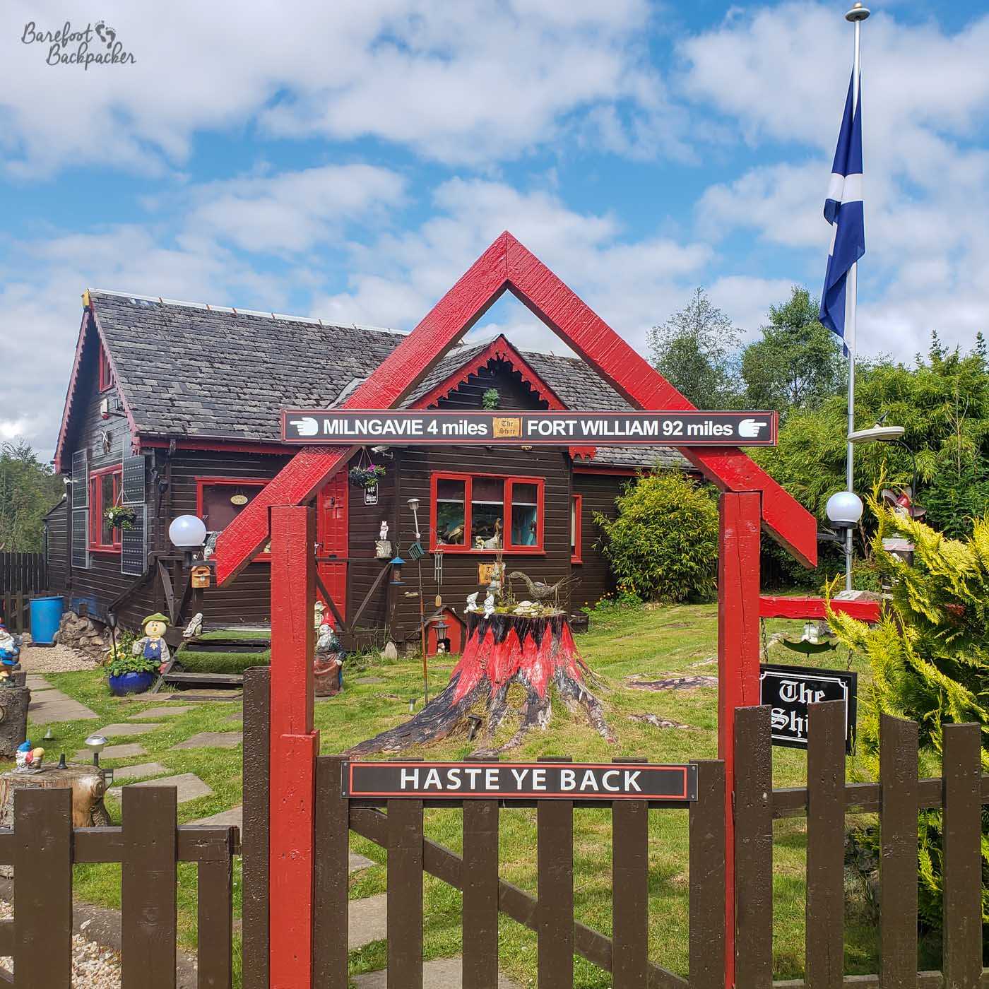 An odd cottage, fronted by a traditional wooden fence with slats,. The cottage has a stone roof but is fronted by what look like wooden panels, like a log cabin. The garden is full of trinkets like gnomes, animal statues, and a tree stump painted to look like fire. Signs on the fence say 'haste ye back', and indicating it's 4 miles to Milngavie and 92 to Fort William. There is a Scotland flag drooping from a flagpole to the right of shot. Lots of signs tell you this cottage is called 'The Shire'.