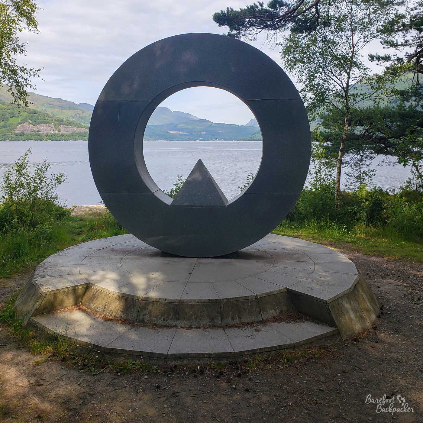 Imagine a metallic ring, wide but not very thick. At the bottom of the ring, covering its thickness, is a small pyramid. Looking beyond the ring is Loch Lomond and the opposite shire in the distance.
