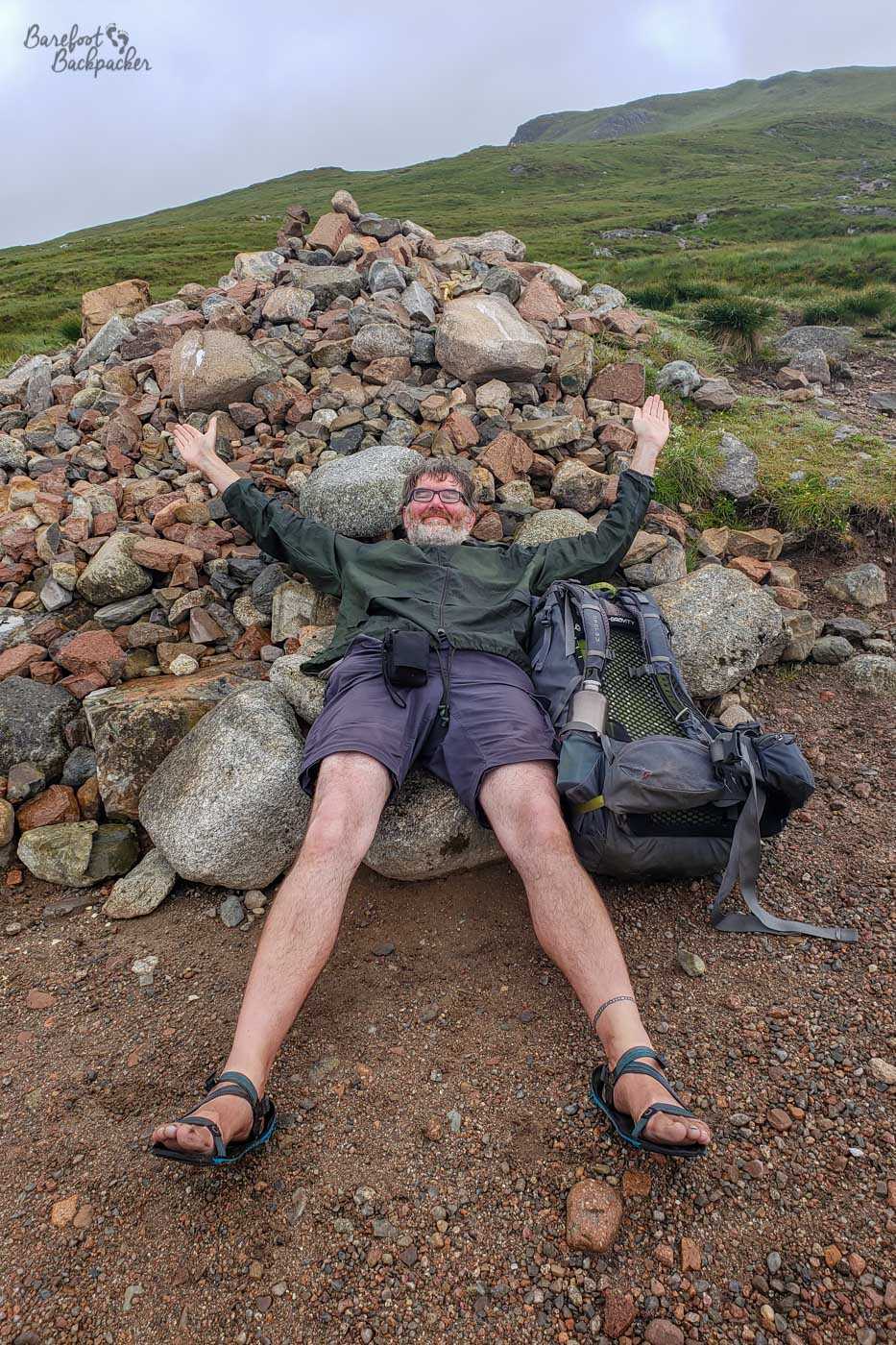 The Barefoot Backpacker is not barefoot. They are wearing minimalist sandals. Ir's at the top of the Devil's Staircase and they are collapsed on a pile of stones, backpack next to them, stretched out in the shape of an X.