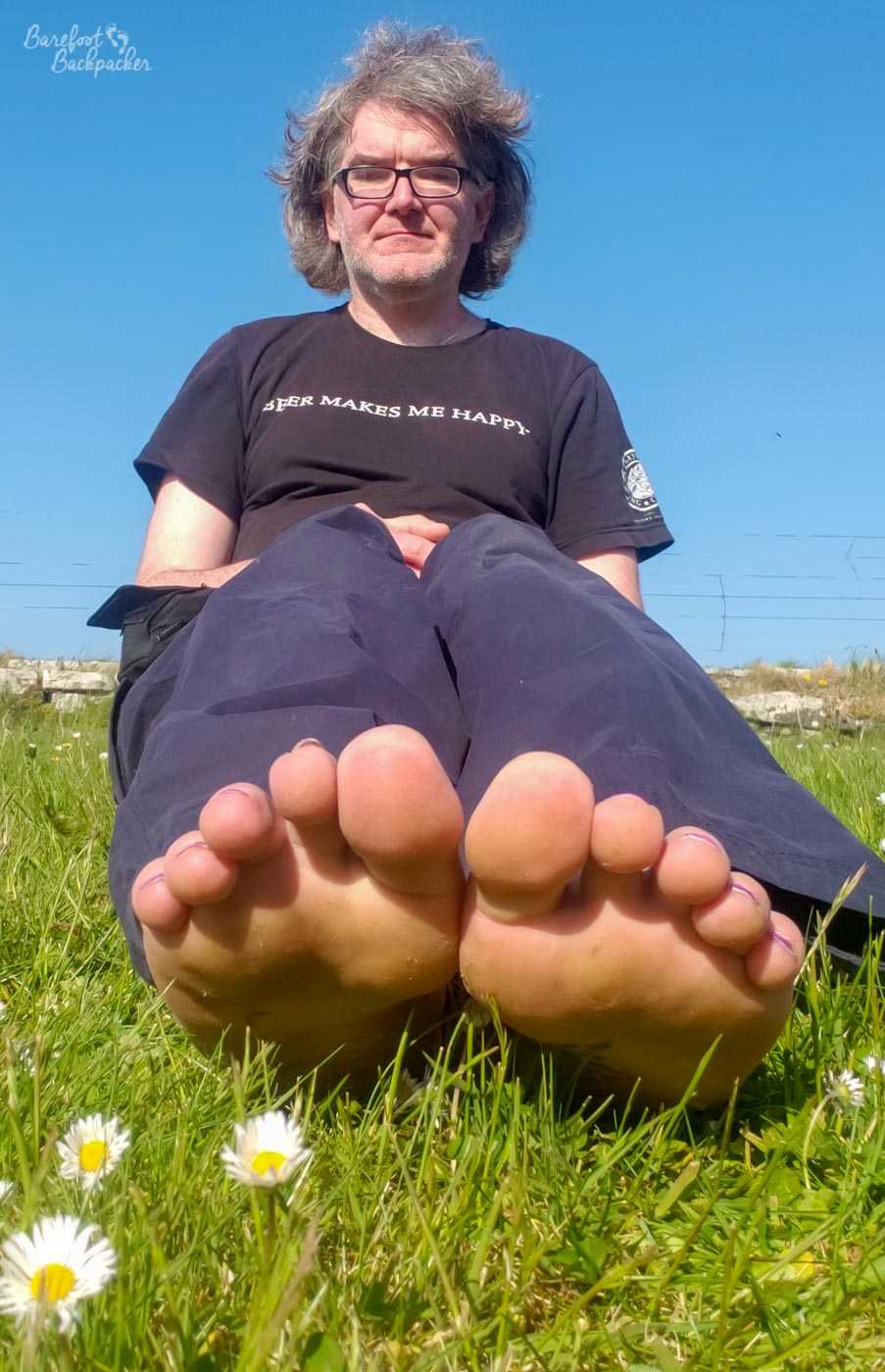 A man (well, an AMAB enby, anyway) is sitting on the grass on a bright sunny day. They are barefoot, and their toes are pointed towards the camera in the foreground, just next to a couple of daisies.