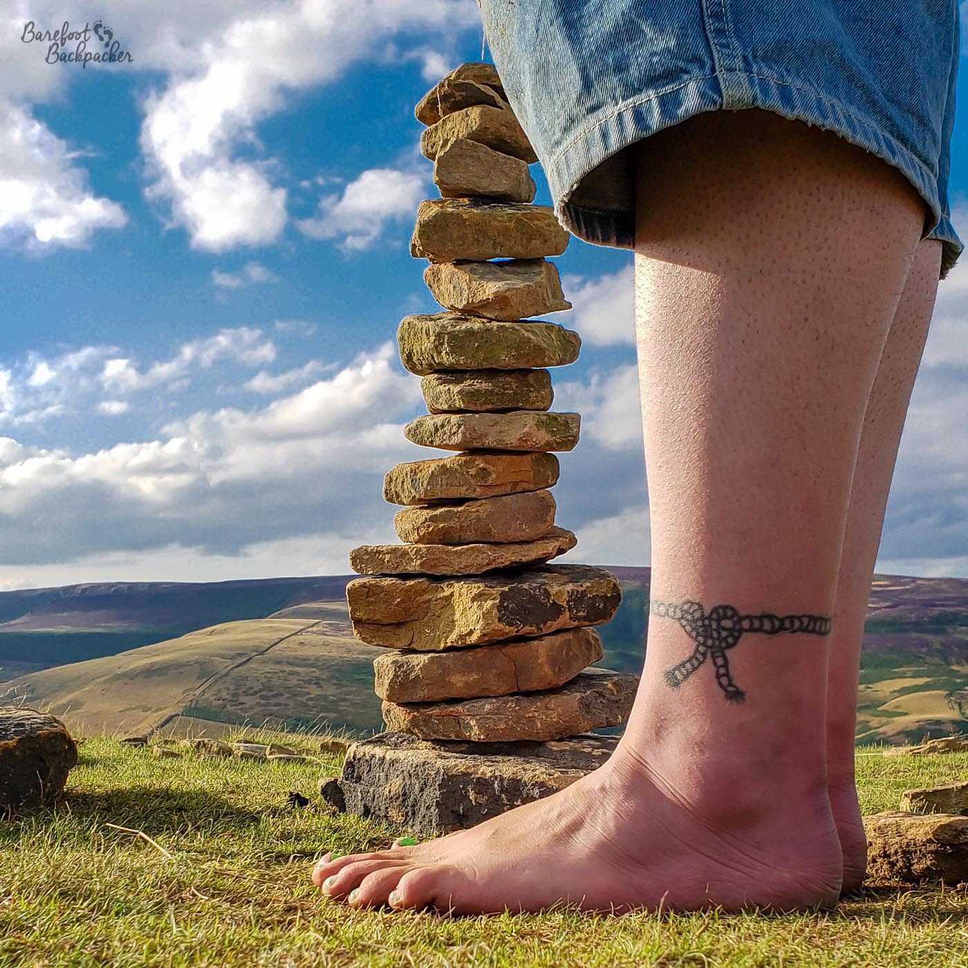 In the background are some rolling hills, and an open sky. The picture is dominated by a small cairn of stones piled up on the grass at the edge of a cliff, and next to and slightly in front, the lower legs and feet of someone are visible standing on the grass. Their denim trousers end just below the knee, and the rest of their legs, and feet, are bare. On the ankle that's visible is a rope motif tattoo.