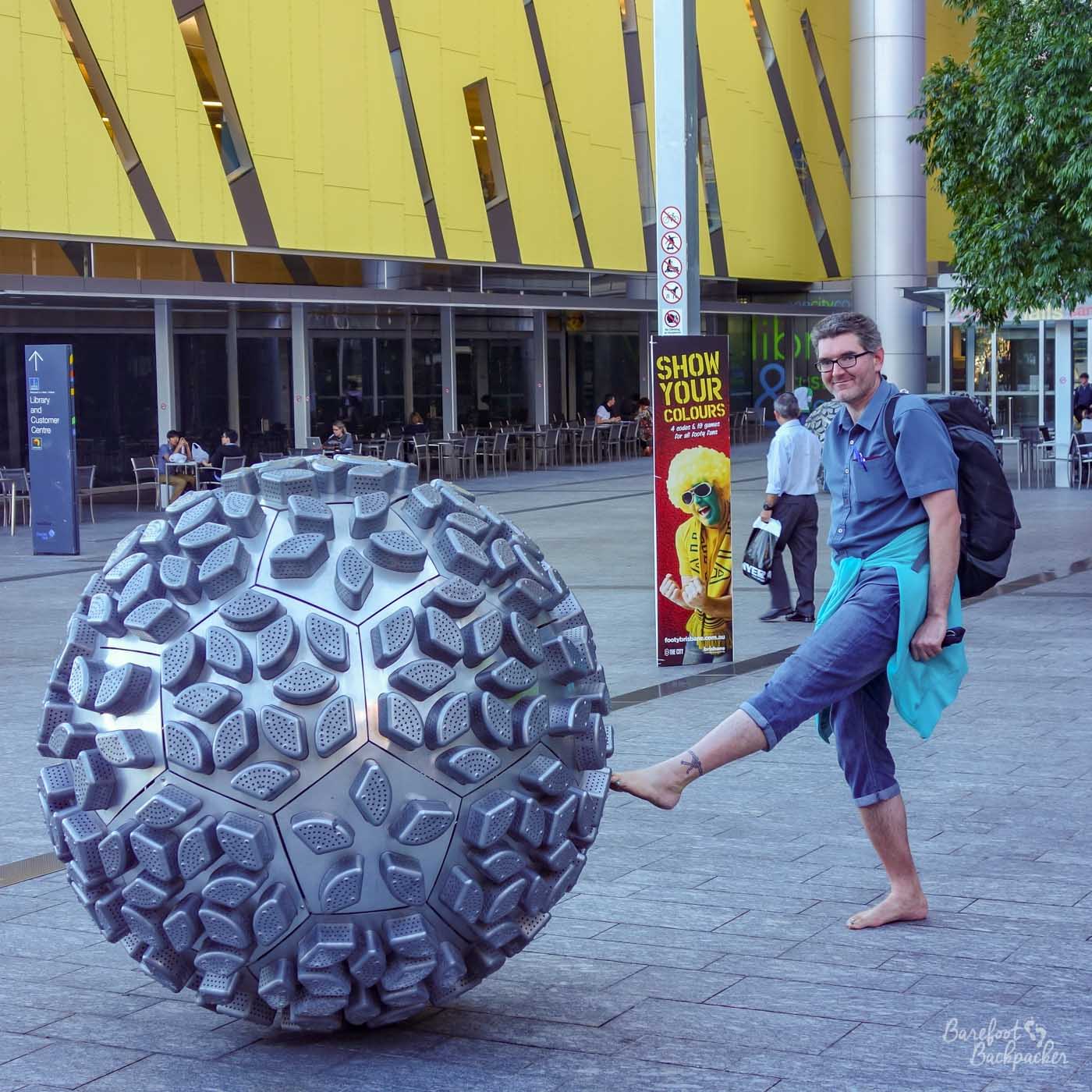A city square, paved area, in front of a building with a row of tables and chairs outside. In the foreground is a large metallic ball, a piece of artwork probably about 1m80 tall, and, as it's a sphere, the same in width. It has segments on it like pentagons and hexagons, and each has a number of sticky-out bits in the shape of wedges. The whole thing has the feel of a very grippy piece of rubber. Anyway. Standing next to the ball, well, pretending to kick it, is a chap with a backpack. The toes of his left foot (he is barefoot) are just touching the ball.