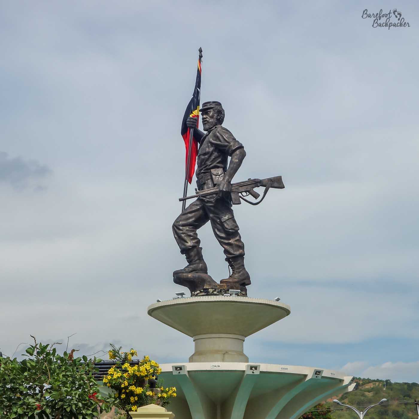 Statue on a circular pillar. He is a soldier. In his left hand he carries a rifle, in his right is the Timor-Leste flag.