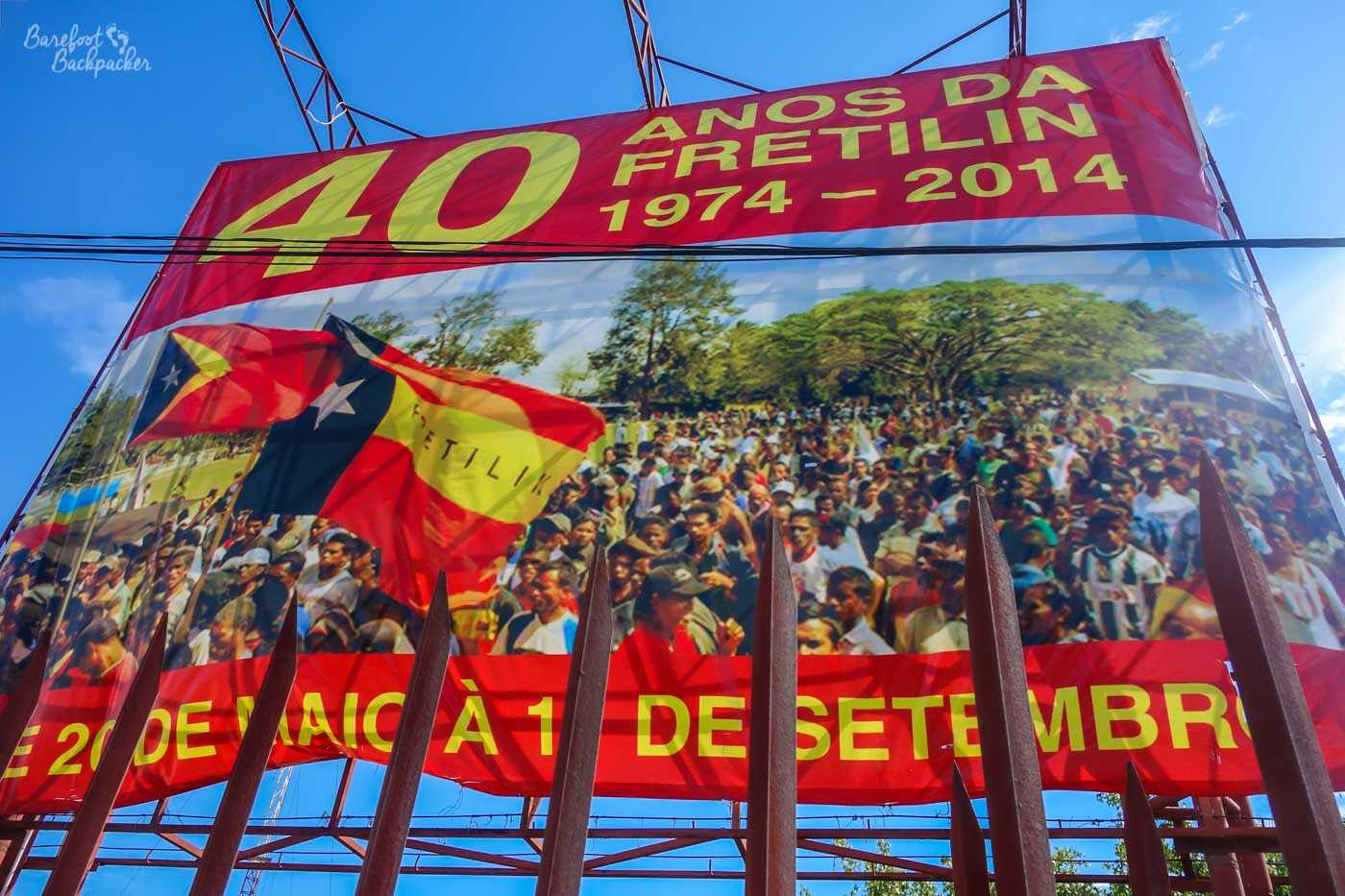 A large poster behind an iron fence, quite high above the ground. It says '40 anos da Fretilin 1974-2014', and shows a large crowd of people with a couple of Timor-Leste flags.