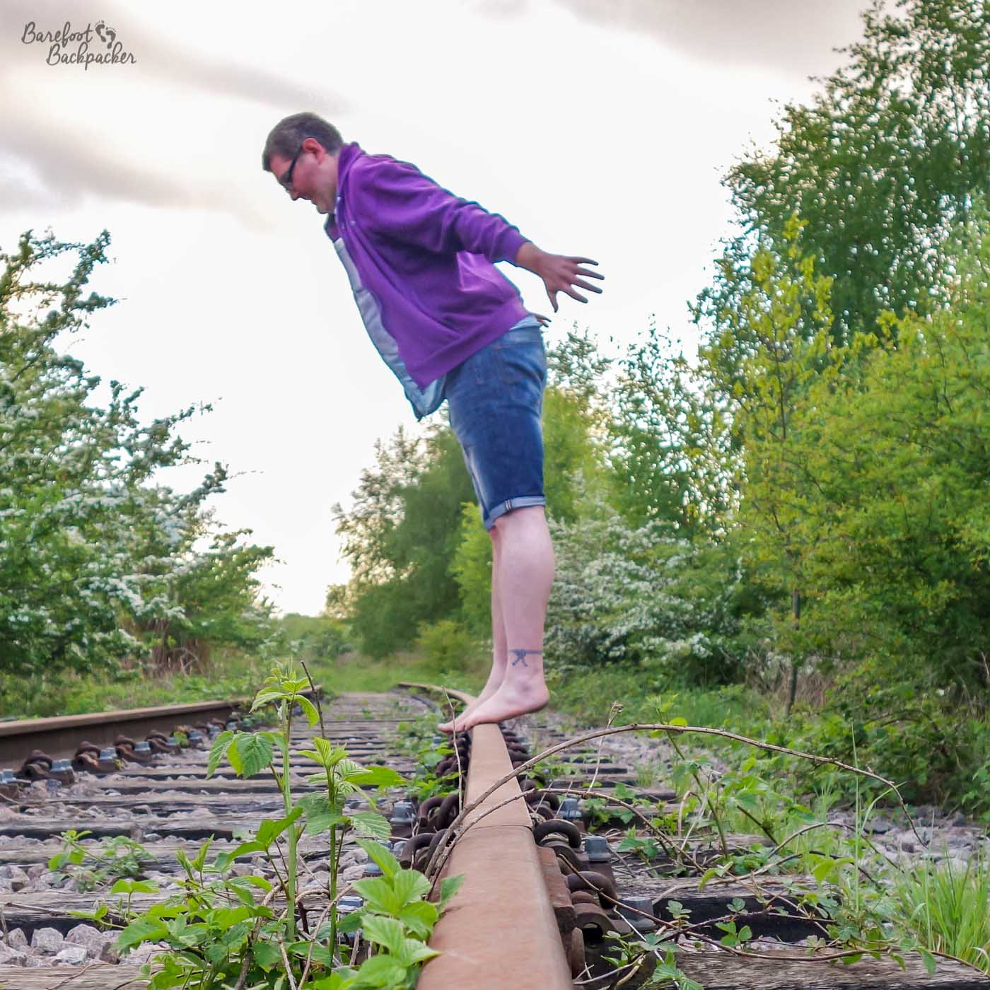 Disused railway line, with vegetation growing across it. An enby is standing on one of the rails, parallel to it and side-on to the camera. They are barefoot, their legs are straight, and they are leaning forward from the waist with arms slightly raised wide, clearly about to fall off.