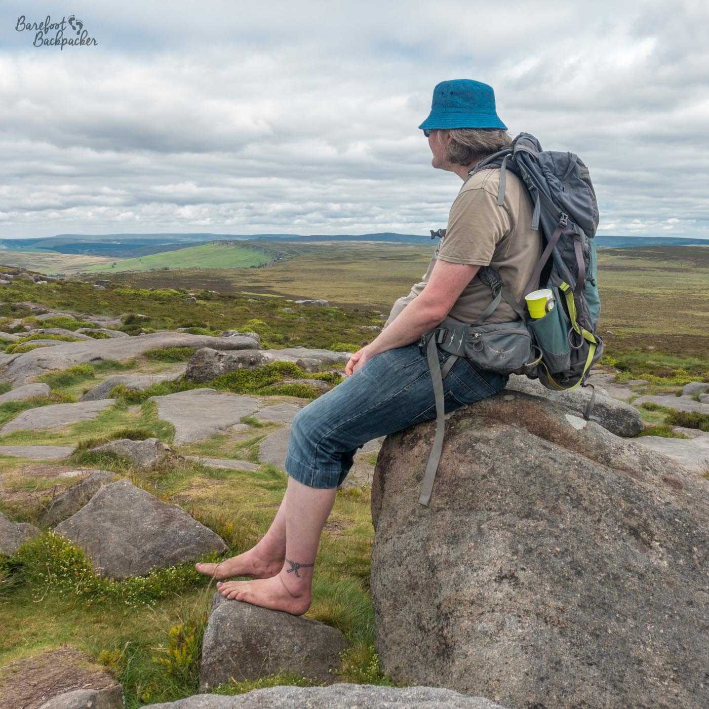 The Barefoot Backpacker sits on a rock in a field of rocks and boulders, staring out over the cliff edge at Stanage Edge in the Peak District. They're barefoot, wearing a hat, t-shirt, and denim shorts that cover the knee, and they're carrying a backpack. In the distance are mossy rounded hills, and the curved edge of the ridge can be seen disappearing into the distance.