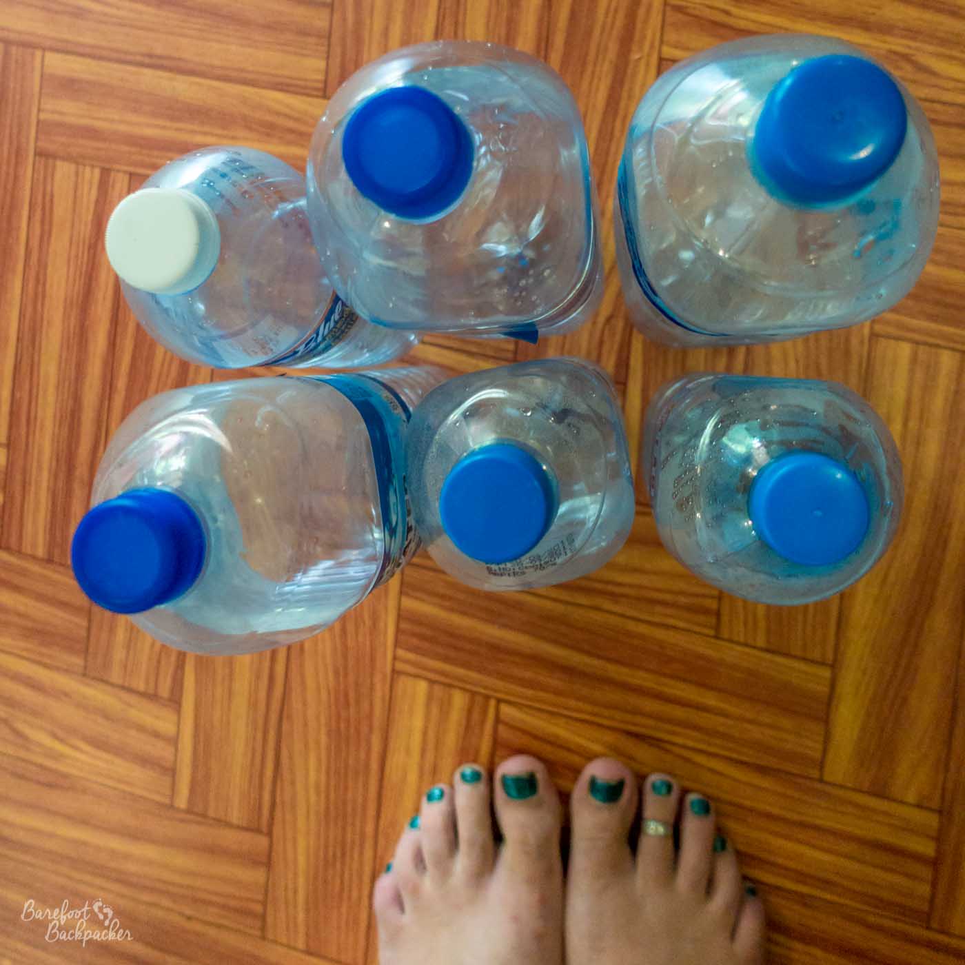 On a parquet floor are six empty plastic single-use water bottles, and below them in shot are the teal-painted toenails and upper parts of two bare feet.