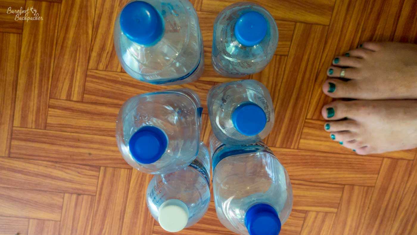 On a parquet floor are six empty plastic single-use water bottles, while to the right of them are the teal-painted toenails and upper parts of two bare feet.