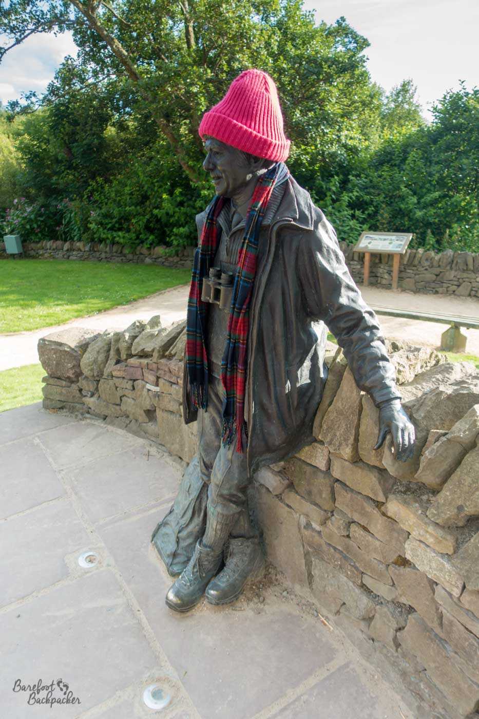 Statue of a man leaning against a wall. He's wearing a jacket, hiking boots, and has binoculars around his neck. At this specific time, he has also been yarn-bombed, and is wearing a knitted hat and a scarf.