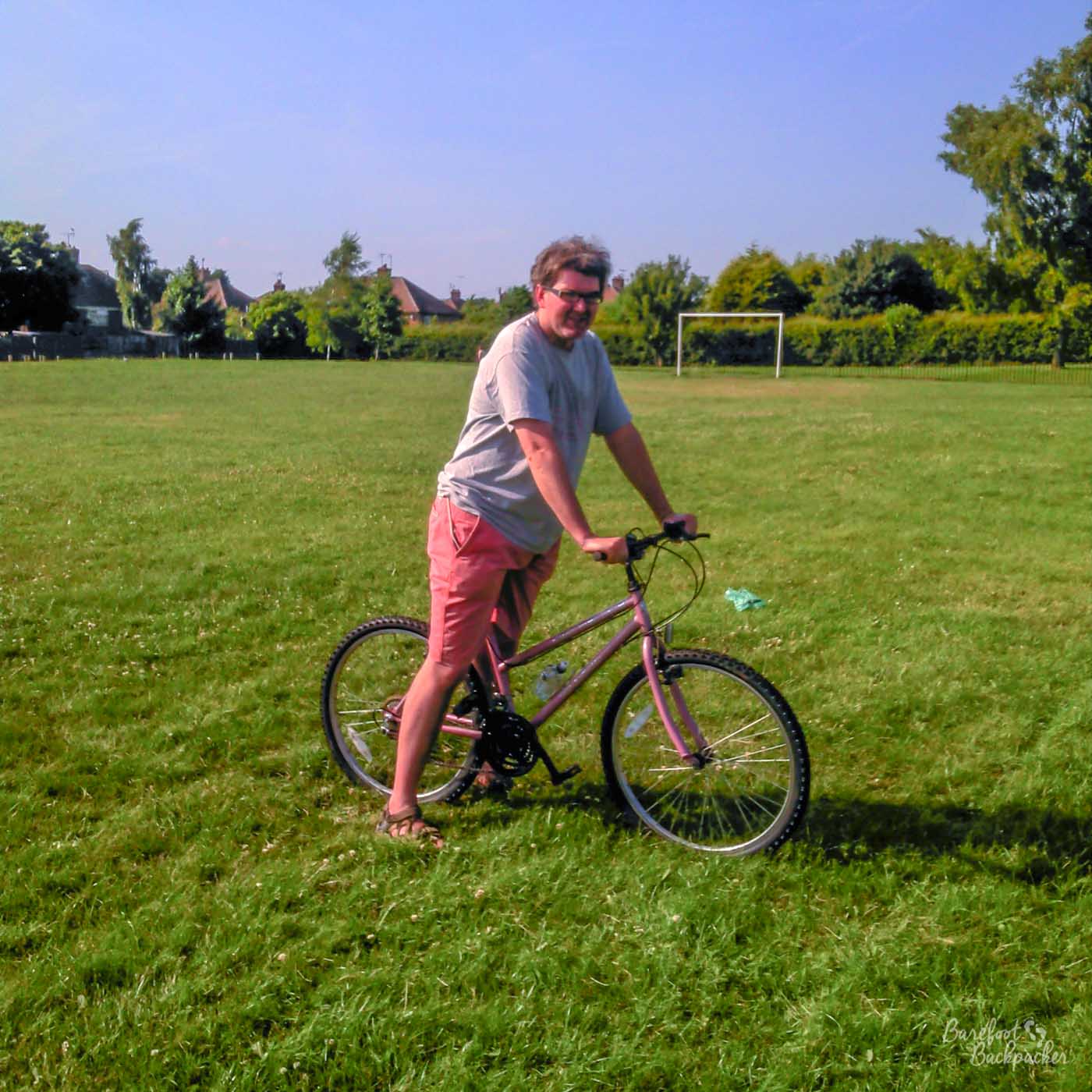A playing field – a goal in front of some trees/bushes in the background, but mostly grass. Someone is kind of standing in the foreground over the saddle of a bicycle, so not riding it but looking like they're about to. The picture is slightly blurry, or at least not particularly sharp/in focus.