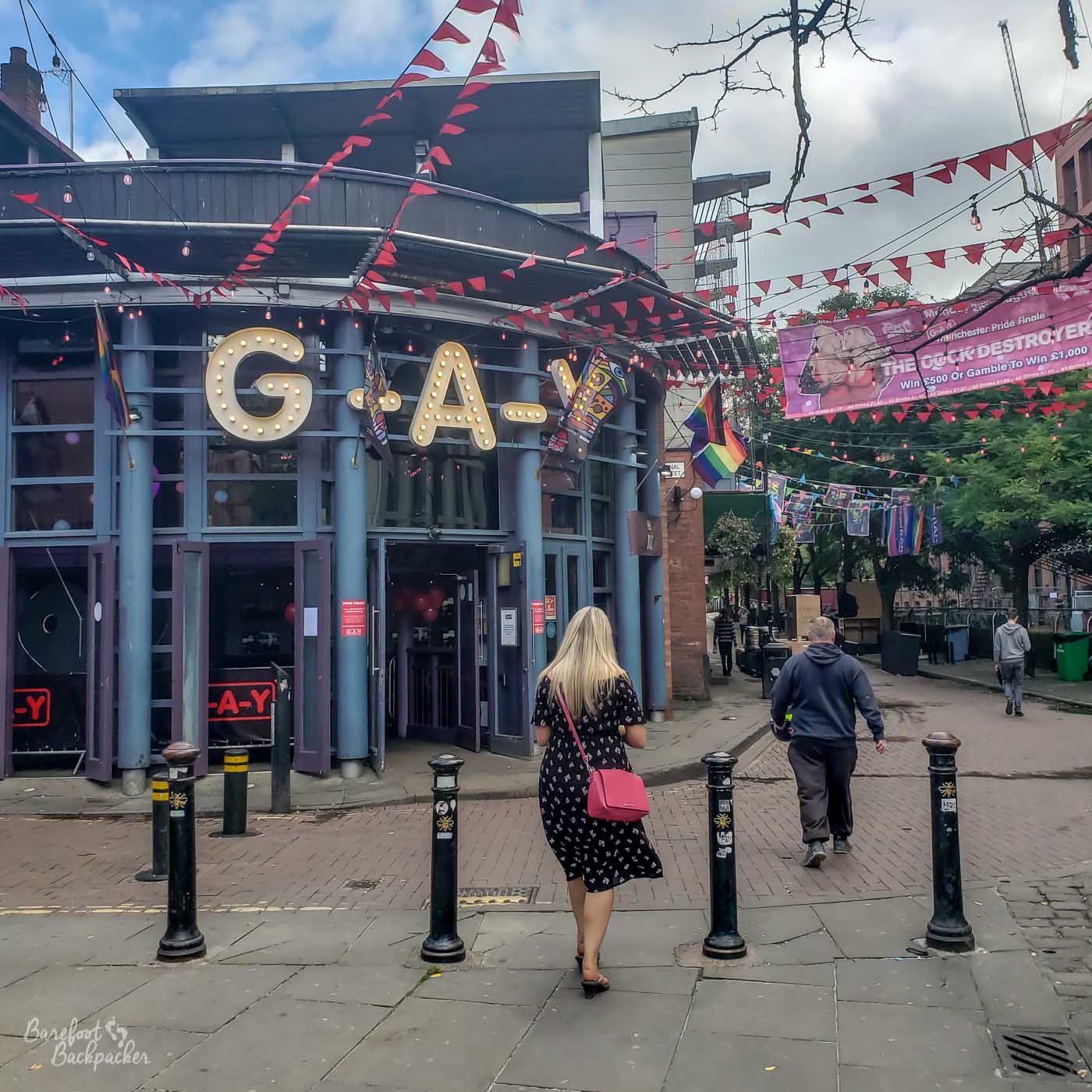 Building with a curved vista with columns in front.. It is a bar called G-A-Y, which are in big yellow LED letters on the front of the building. It is at a junction of pedestrianised roads – the road  to the right of the building is bedecked in bunting of rainbow colours. It's Pride Week.