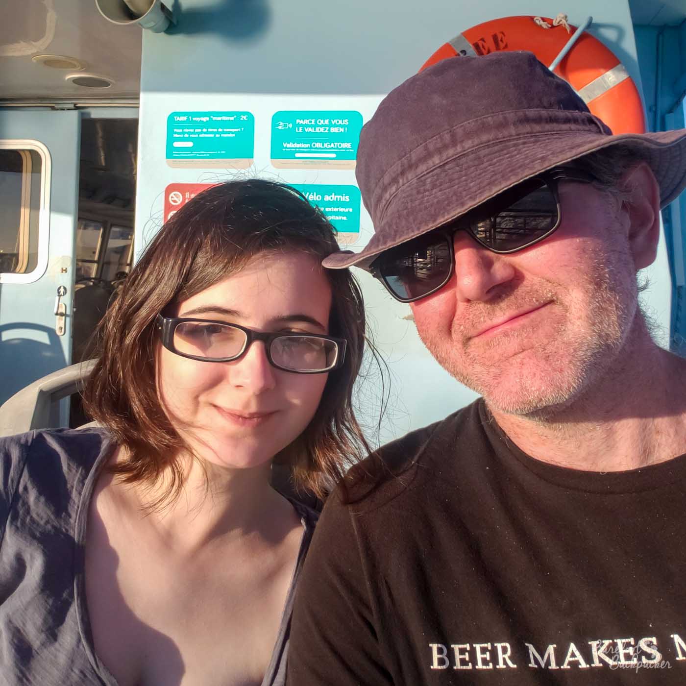 two people on the deck of a small ferry. They are both wearing glasses. One is female-presenting with shoulder-length hair, and is smiling. The other is male-presenting, wearing a hat and has a bit of facial stubble.