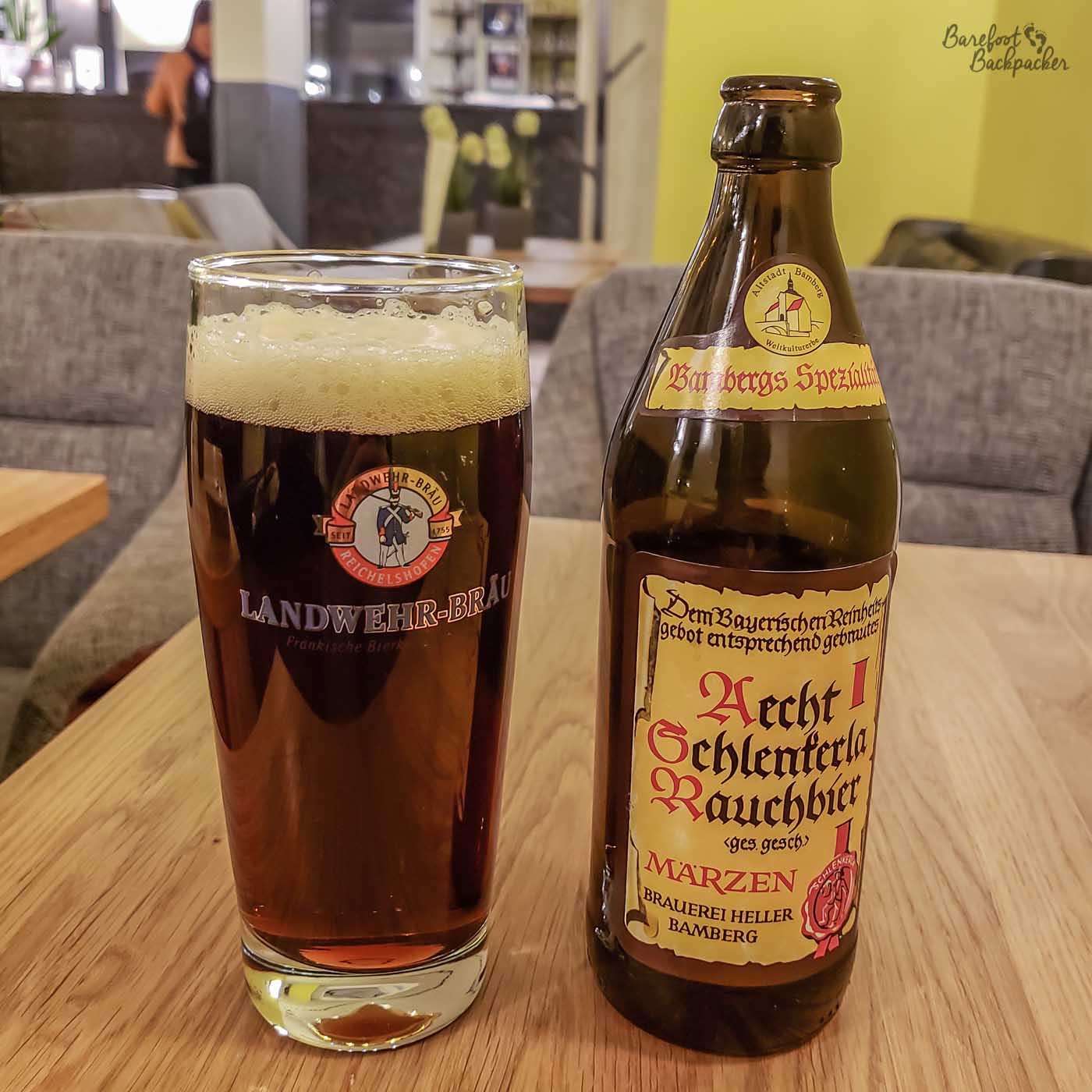 A bottle of beer stands on a table. To the left of it is a glass with the beer in. The beer is 'Aecht Schlenkerla Rauchbier' from Bamberg Brewery, Bavaria. The writing on the bottle is in an old-fashioned type script, as if it were written with a fountain pen or quill.