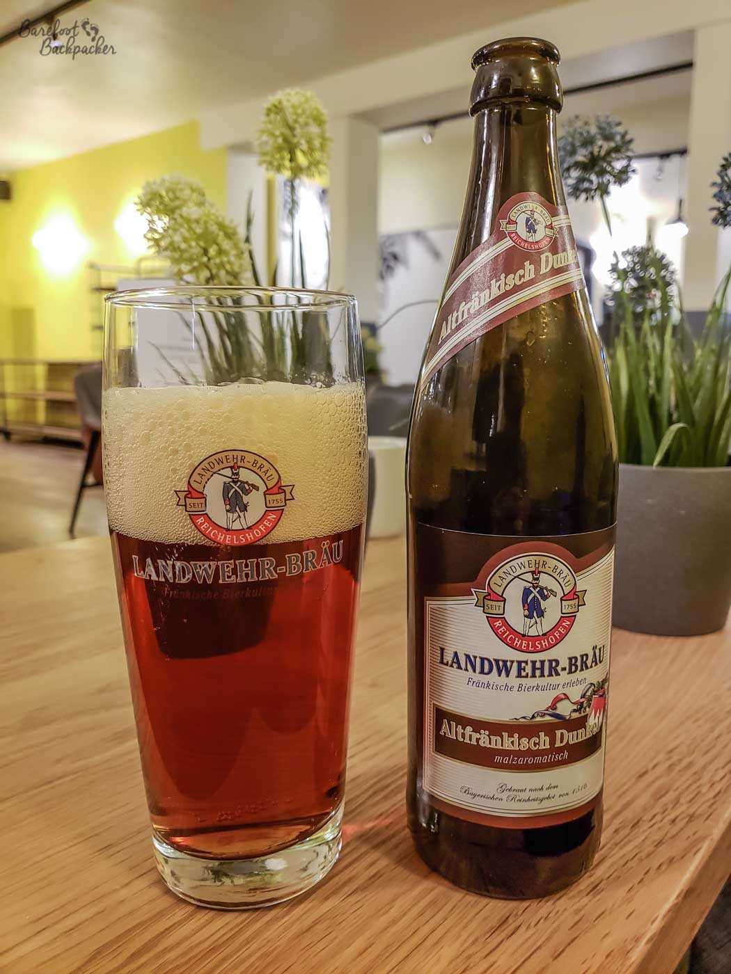 A bottle of beer labelled as 'Landwehr-Brau' is on a table next to a glass, also branded the same. The beer is more of an amber shade. At the bottom of the label on the bottle are the words 'gebraut nach dem Bayerischen Rheinheitsgebot von 1516' – translated to English, it means 'brewed after the Bavarian Rheinheitsgebot of 1516'.