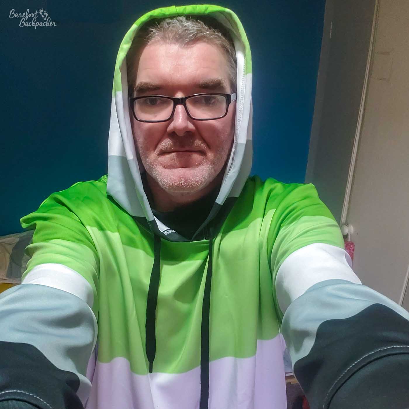 A person is sat in a room with dark walls. They are wearing a hoodie that covers their head, so only their face is visible. They are wearing glasses. The hoodie is striped in the colours of the aromantic pride flag (from top to bottom – dark green, light green, white, grey, black).