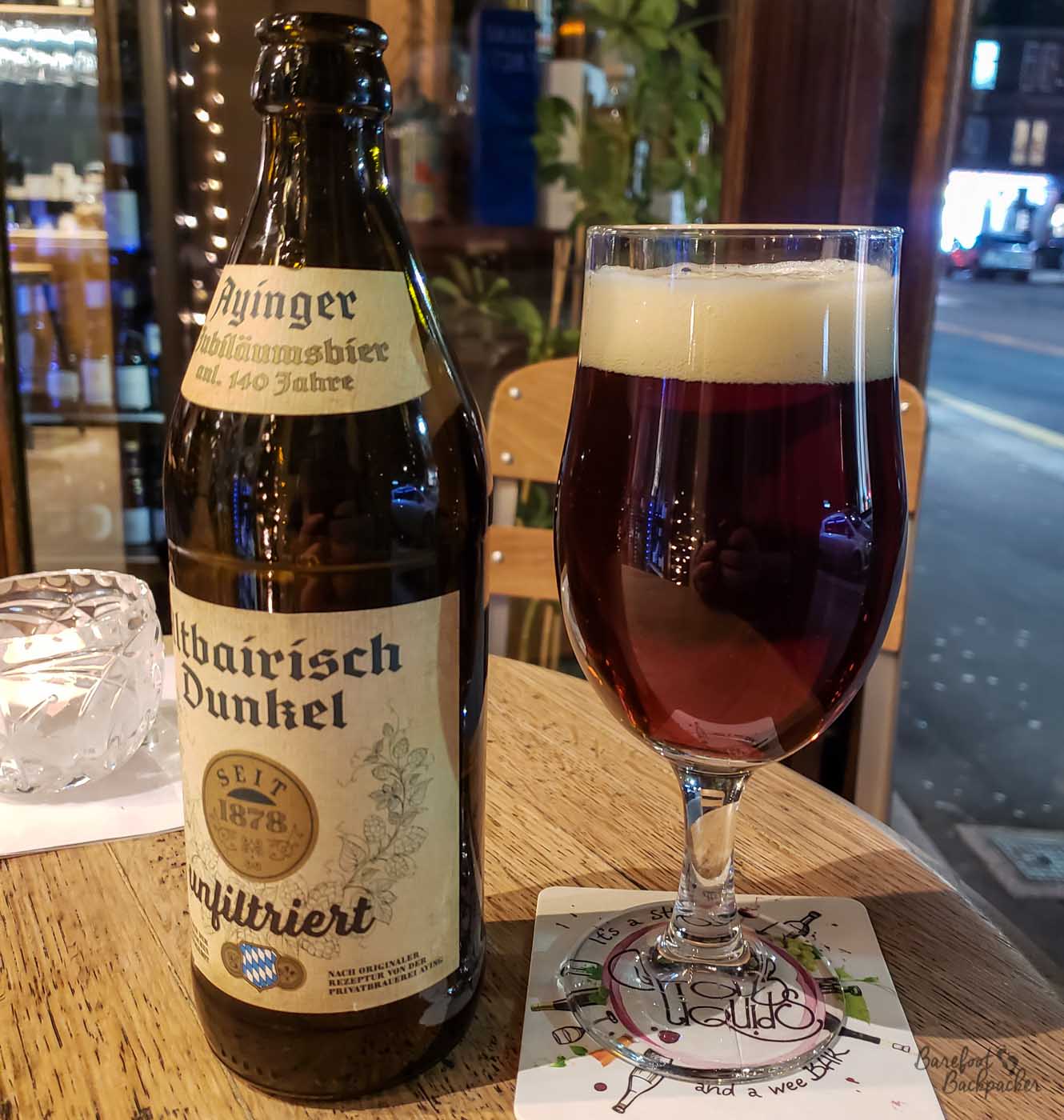 A bottle of beer stands on a table. To the right of it is a glass with the beer in. The beer is 'Ayinger Altbarisch Dunkel'.