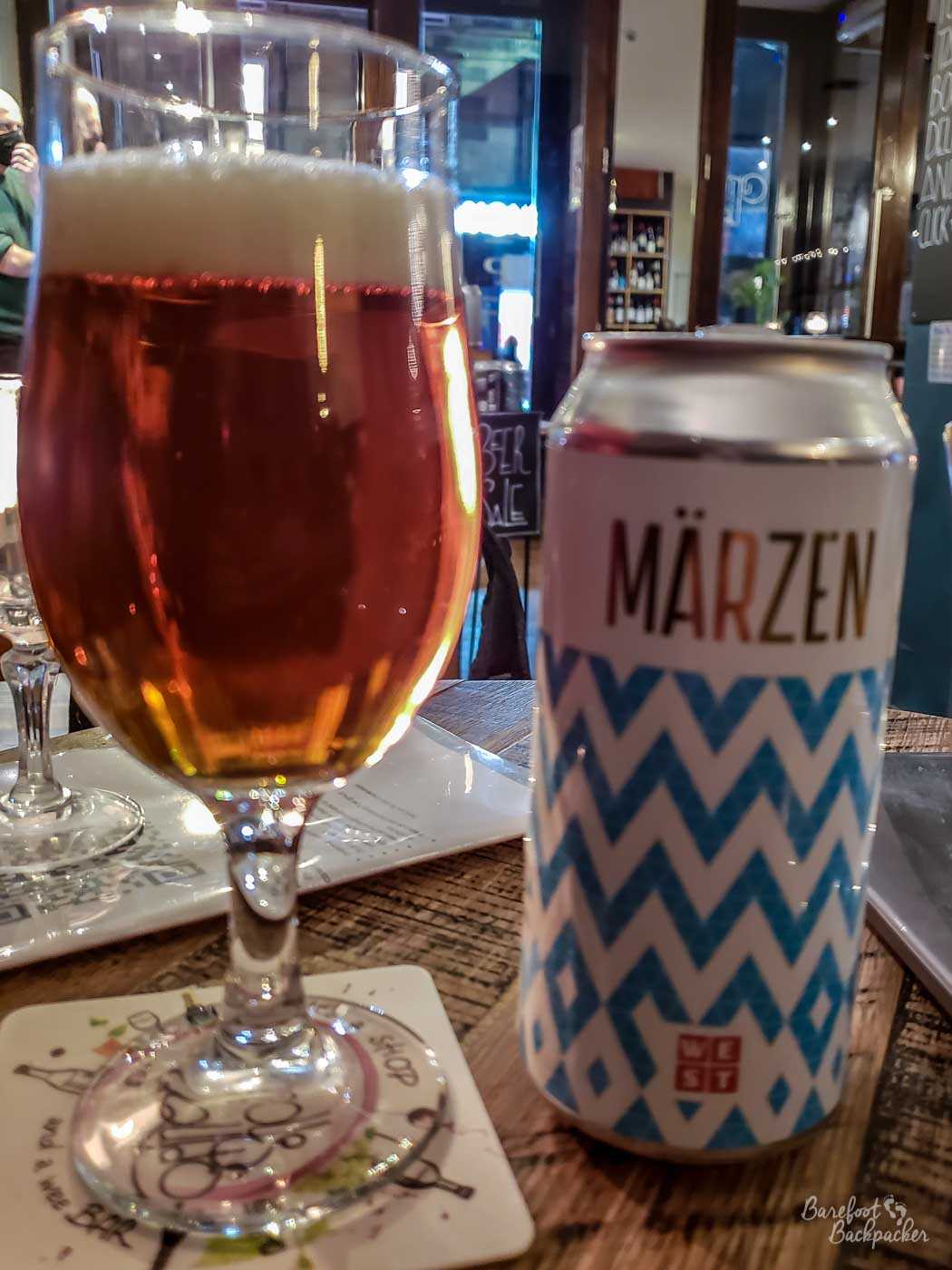 A can of beer stands on a table. To the left of it is a glass with the beer in. The beer is 'Marzen', from West Brewery. The can is decorated in a zig-zag pattern.
