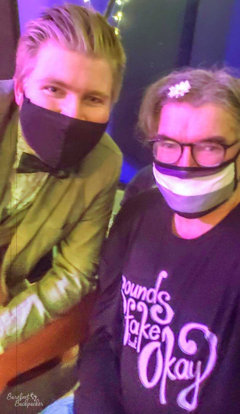 Two people in shot. Both in asexual pride facemasks. Pic is taken inside a dark club or bar venue. The person on the left is wearing a leathery coat. The person on right has a daisy hair clip and a t-short that says 'sounds fake but okay' in retro-styled lettering.