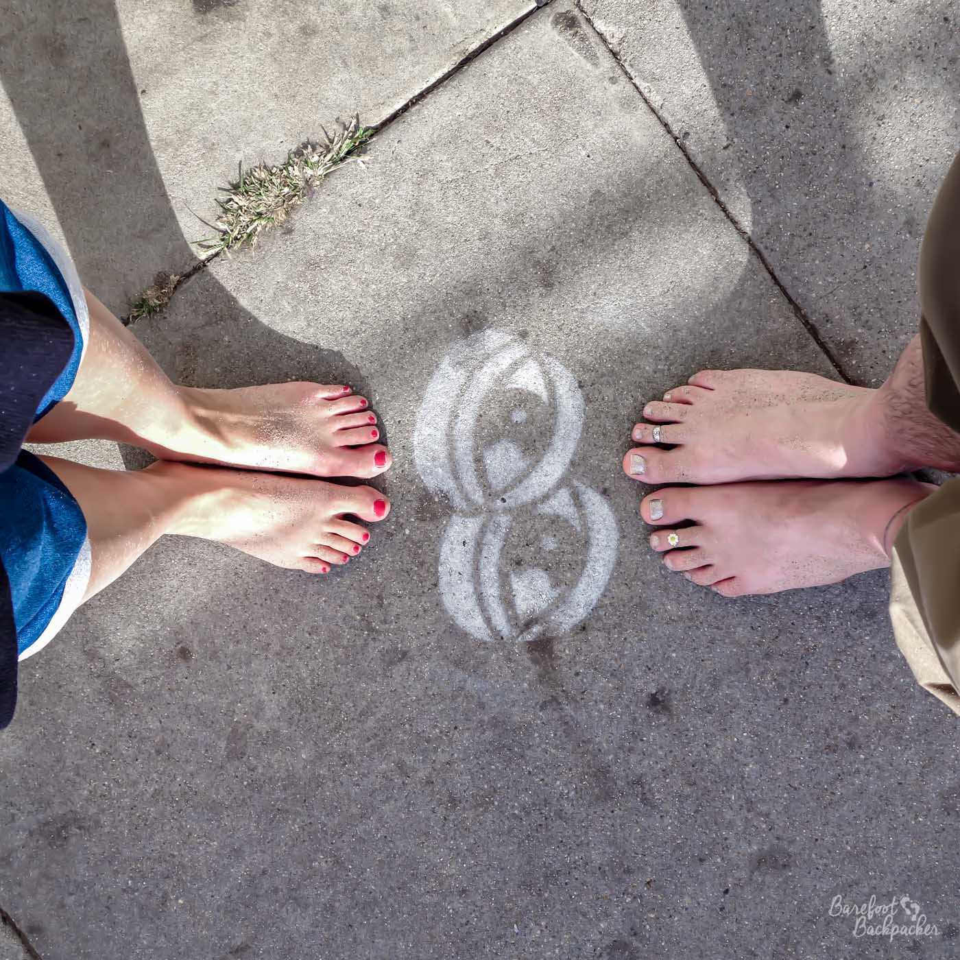 A shot from above looking down at the pavement. In the centre of the shot are a pair of eyes, drawn in white chalk. On either side of the eye art are a pair of feet, bare, the one on the left with red nail varnish, the one on the right with silver nail varnish.